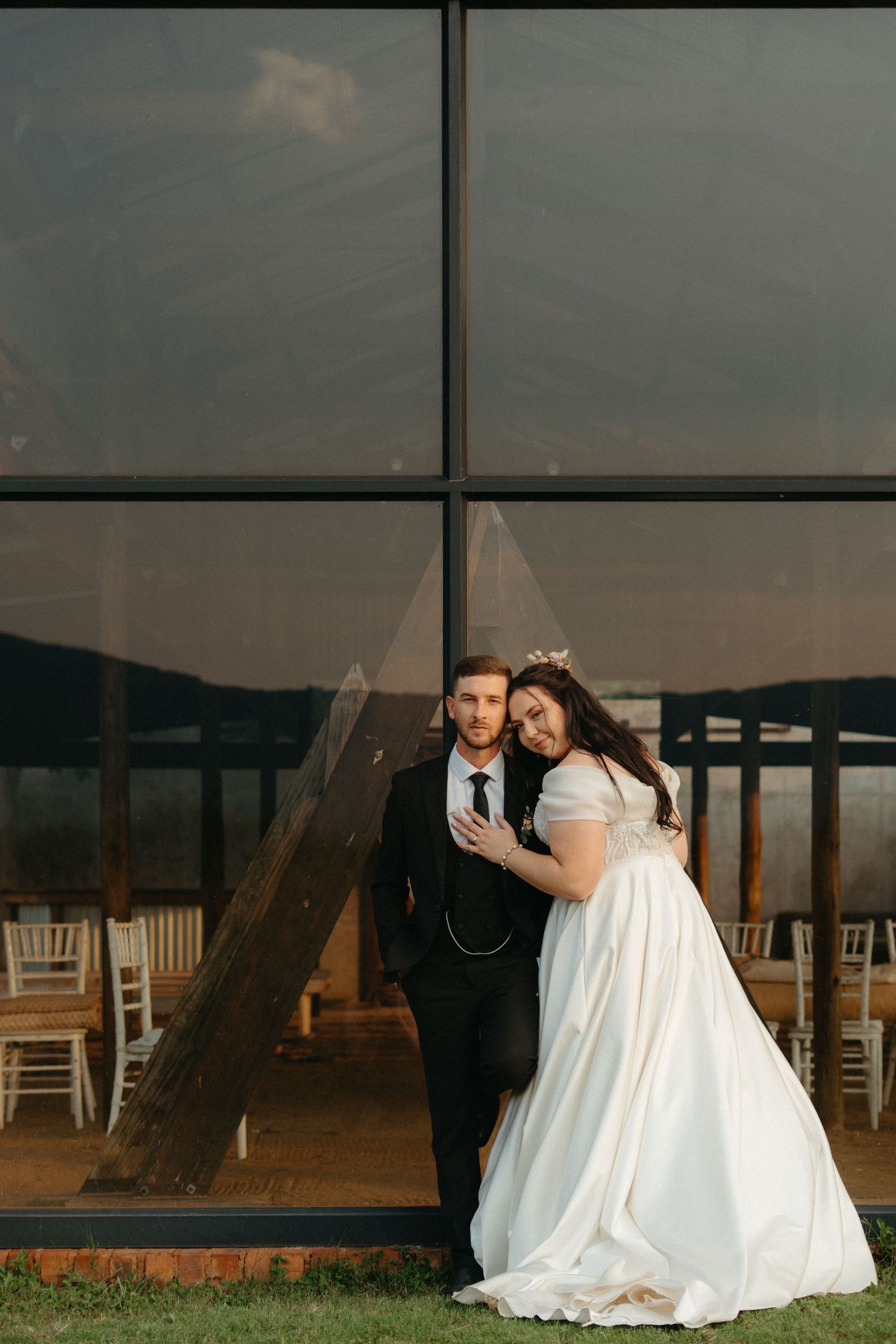 Couple in elegant wedding attire pose against reflective glass panels at sunset.