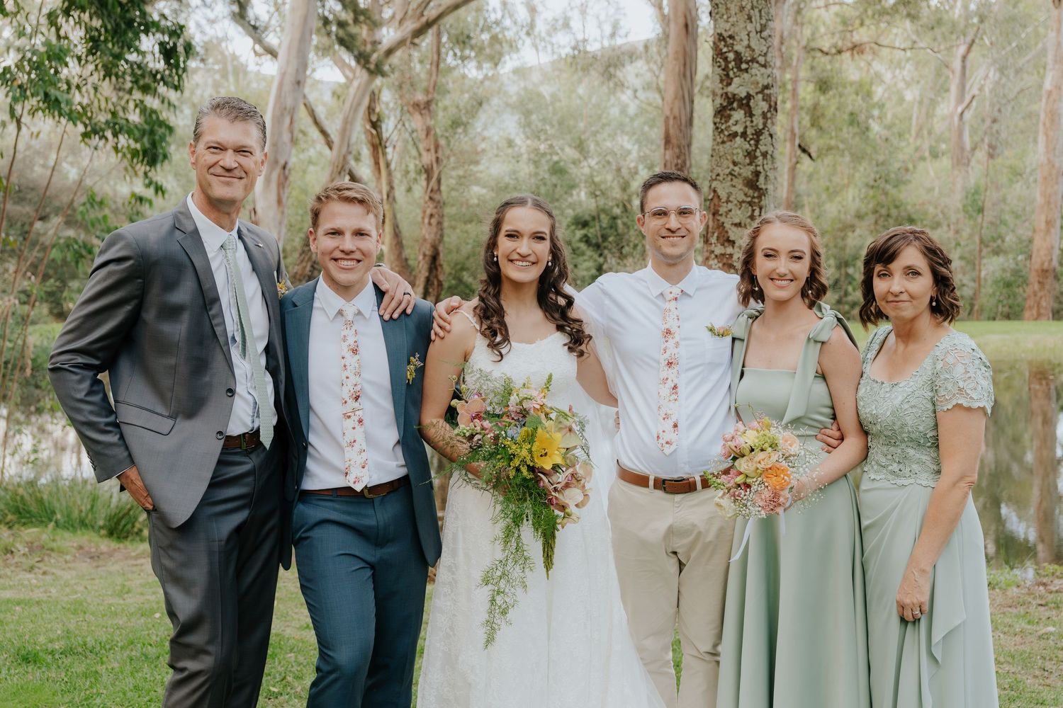 Wedding party in sage green and neutral attire poses outdoors with yellow bouquet against natural woodland backdrop.