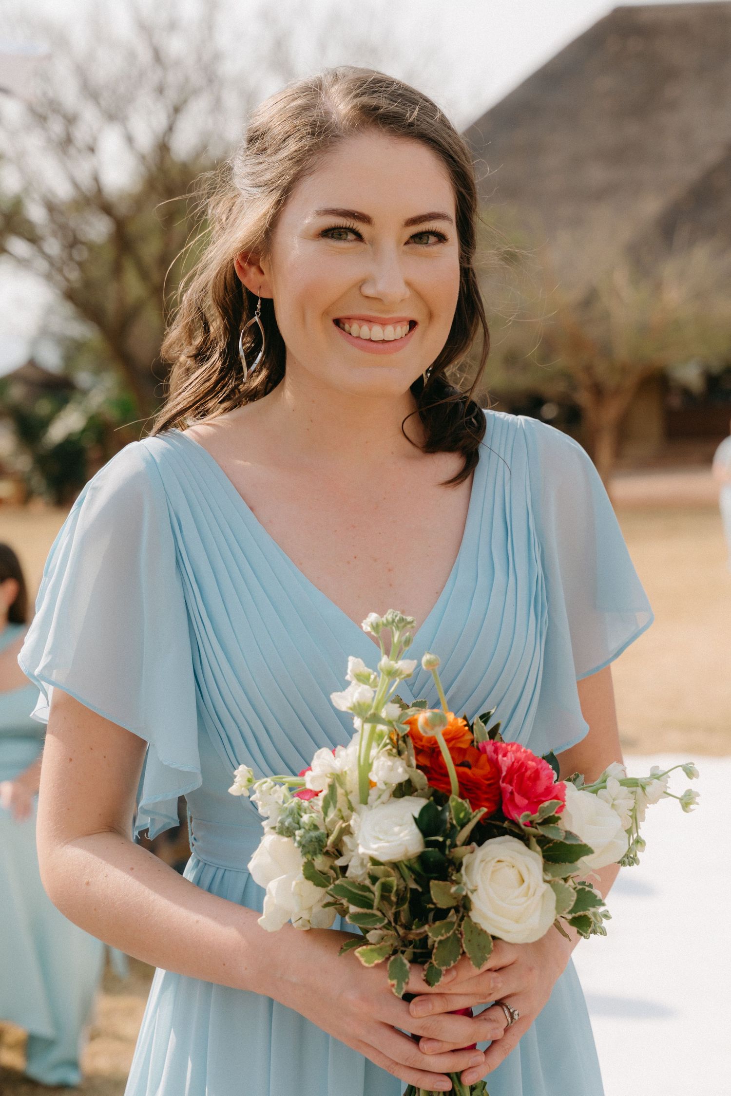 Bridesmaid in flowing light blue dress holds a white and red floral bouquet against natural backdrop.