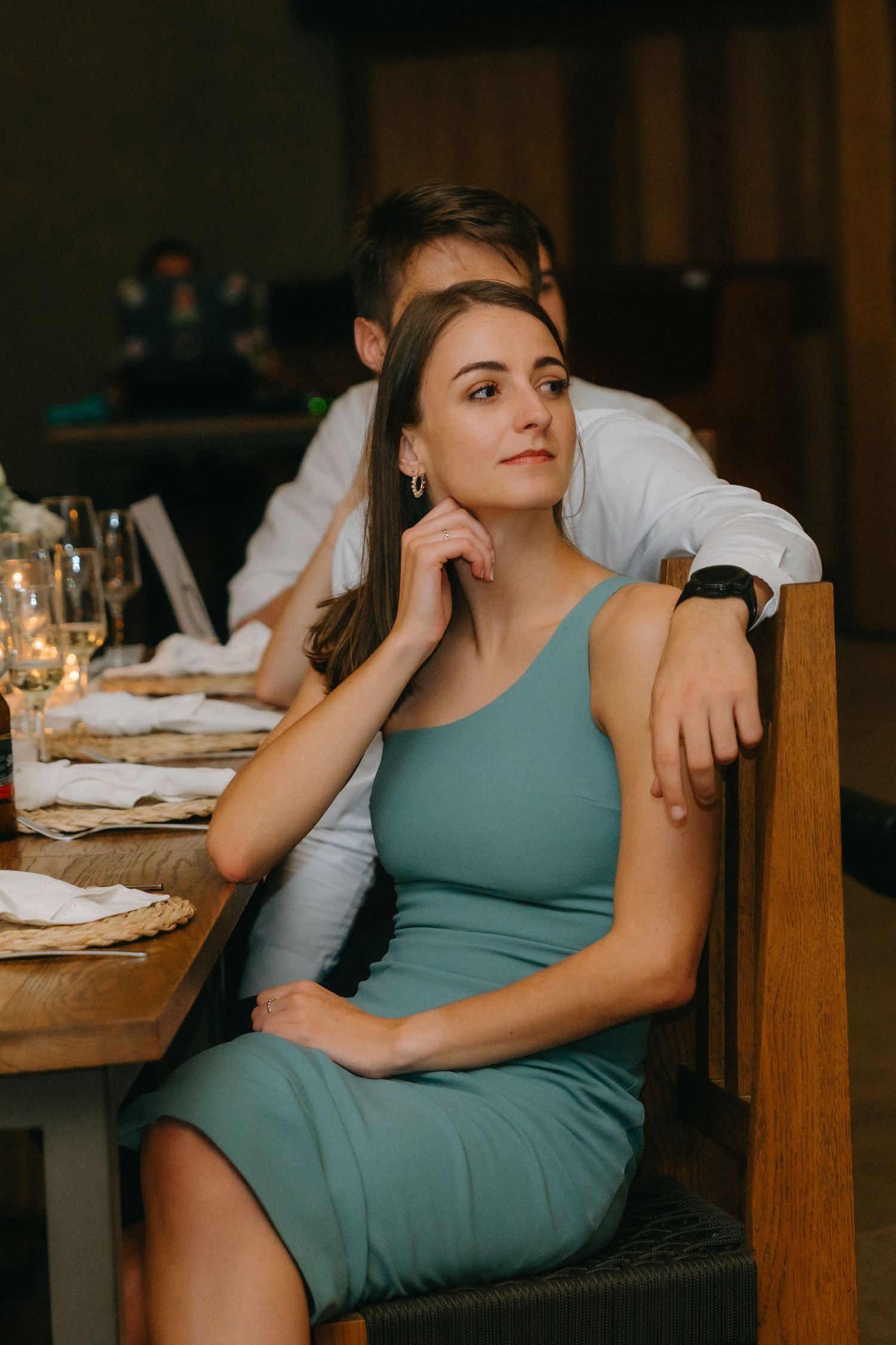 A romantic restaurant scene with someone in a teal fitted dress sitting at an elegant table setting in warm lighting.