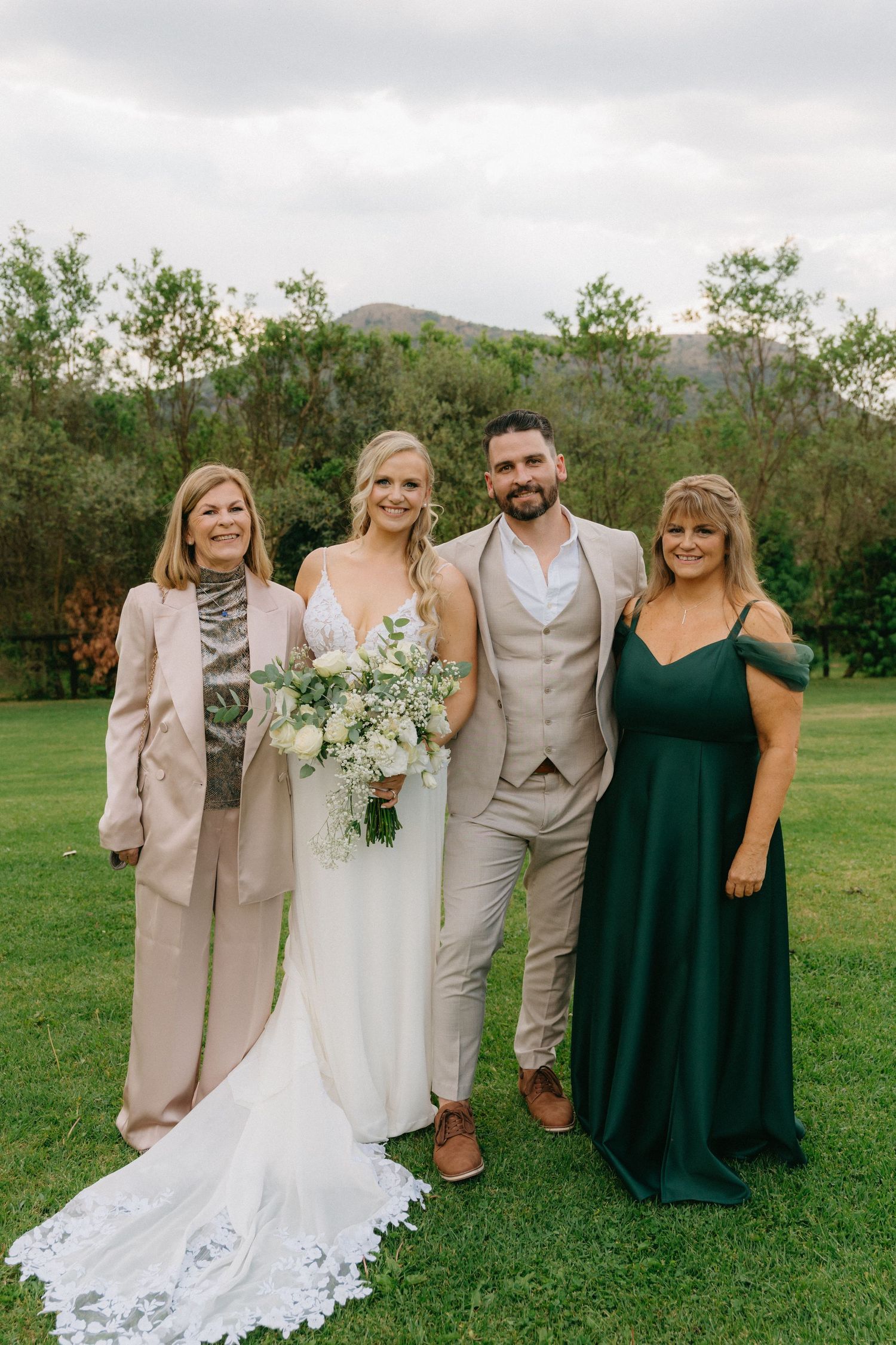 Wedding party poses outdoors in sage green dresses and beige suits against a mountain backdrop.