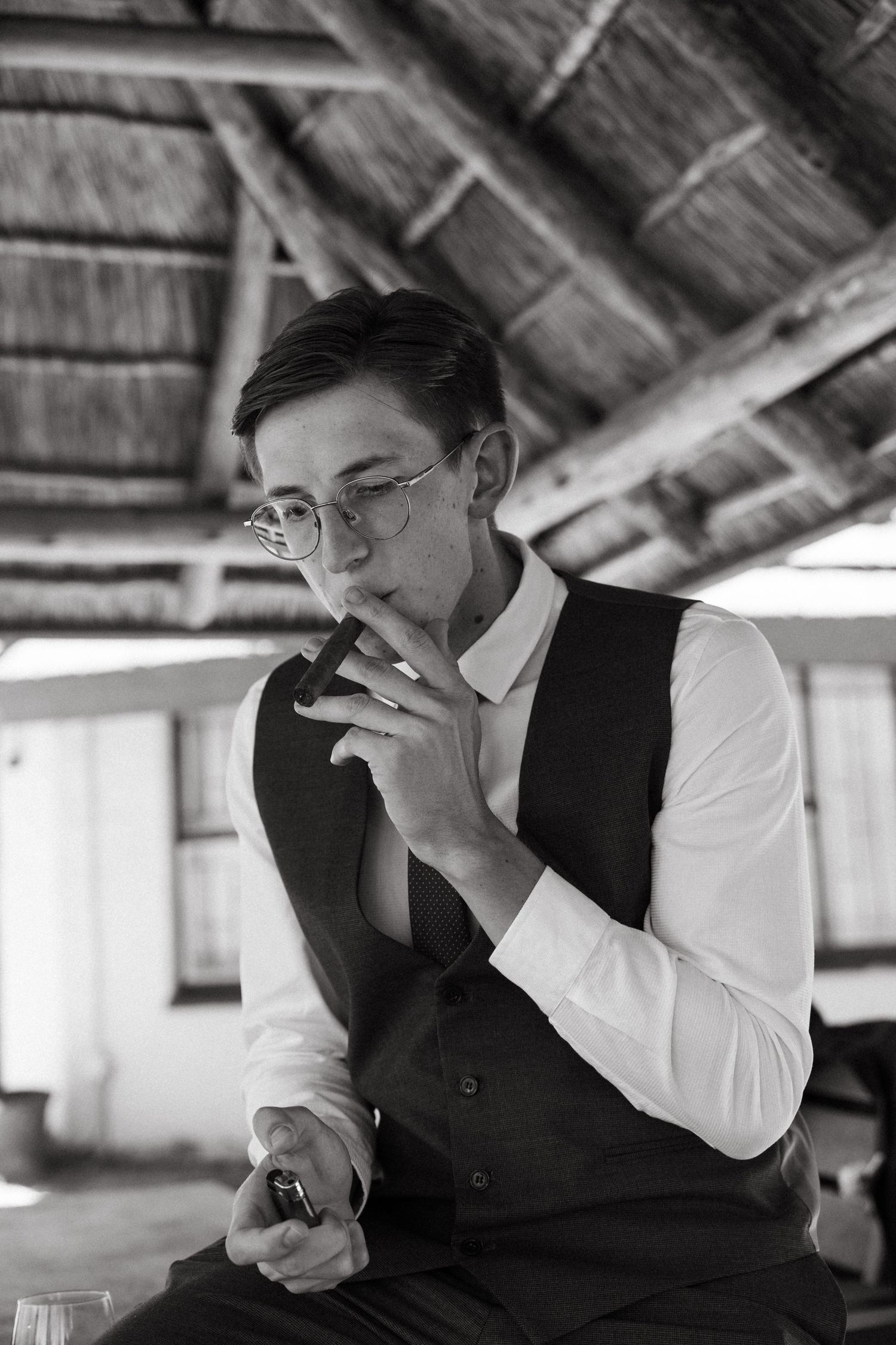 Black and white portrait of a person wearing glasses, vest and white shirt, smoking in a rustic attic setting.