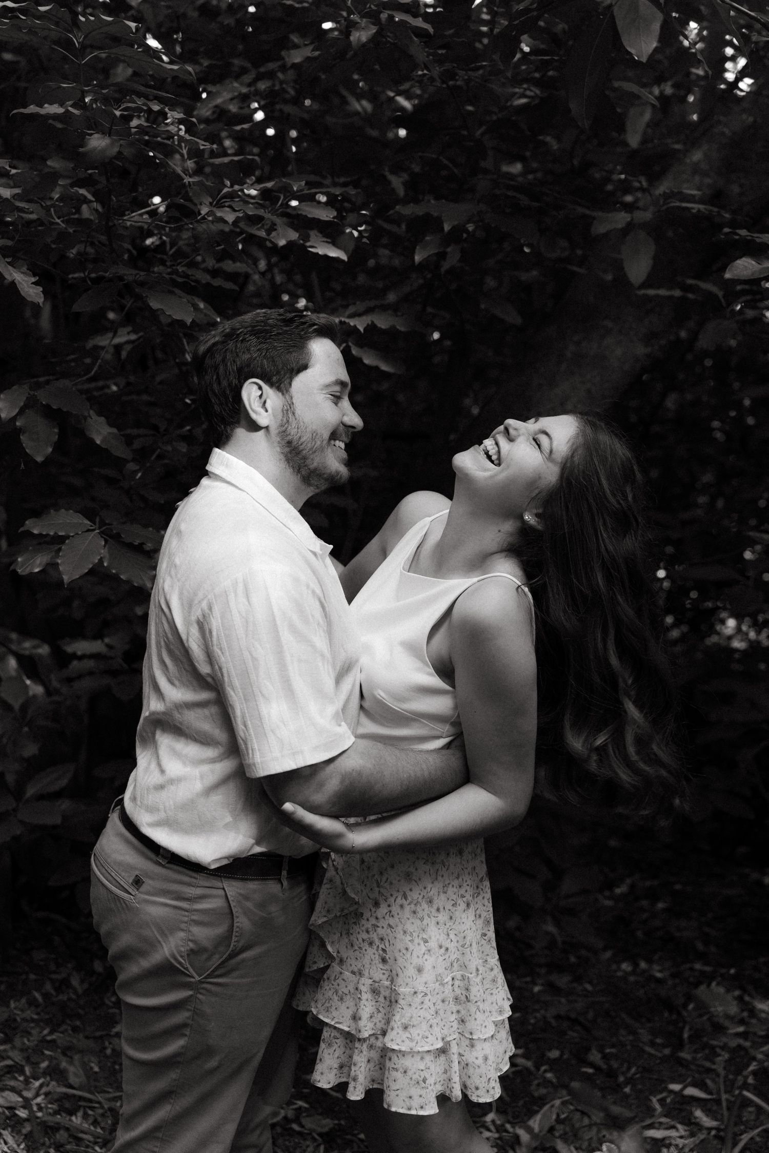 Black and white photo of a laughing couple embracing outdoors among lush green foliage during an engagement session in Pretoria