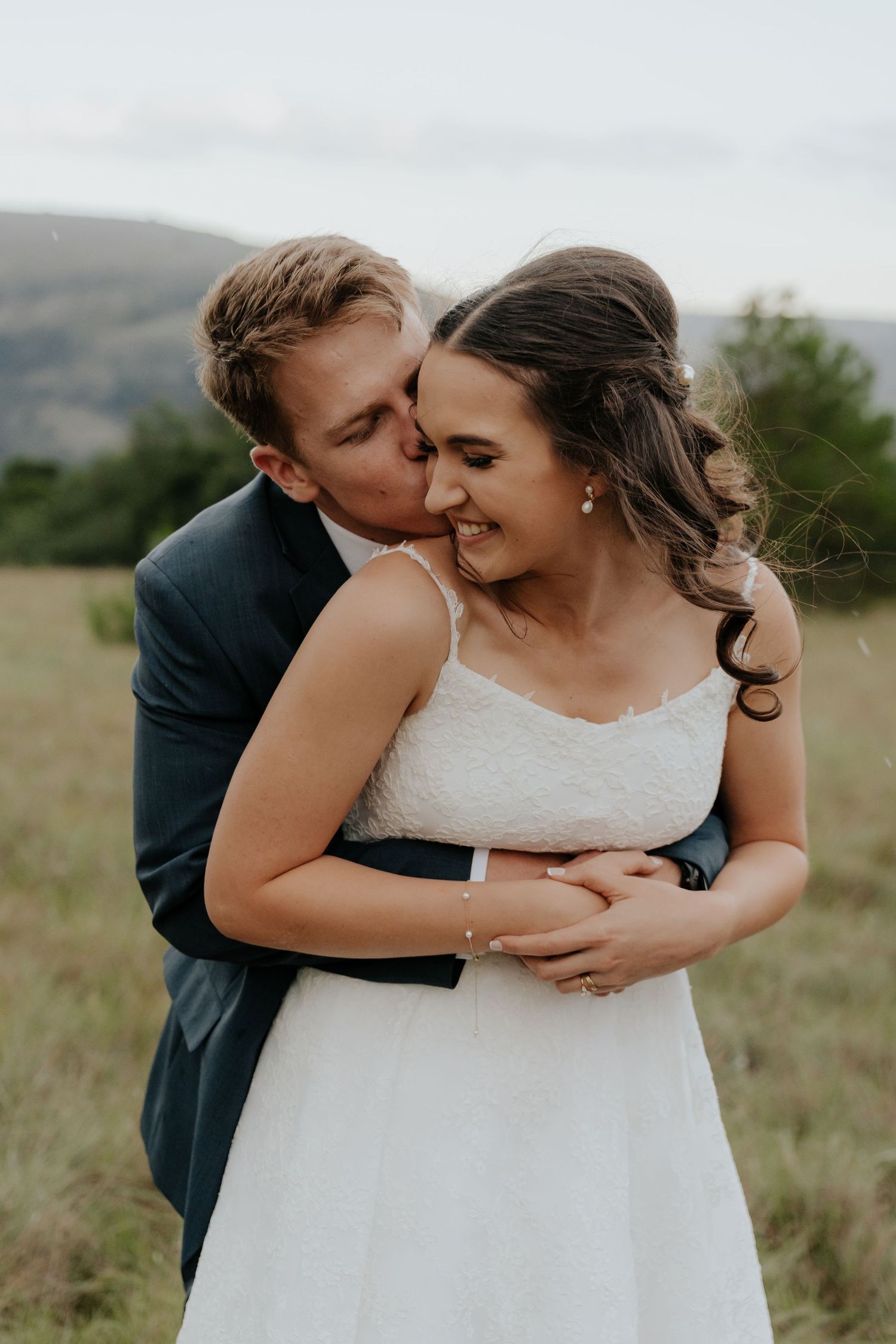 A couple shares intimate moments in a grassy field, with the bride wearing a white lace dress and groom in dark suit.