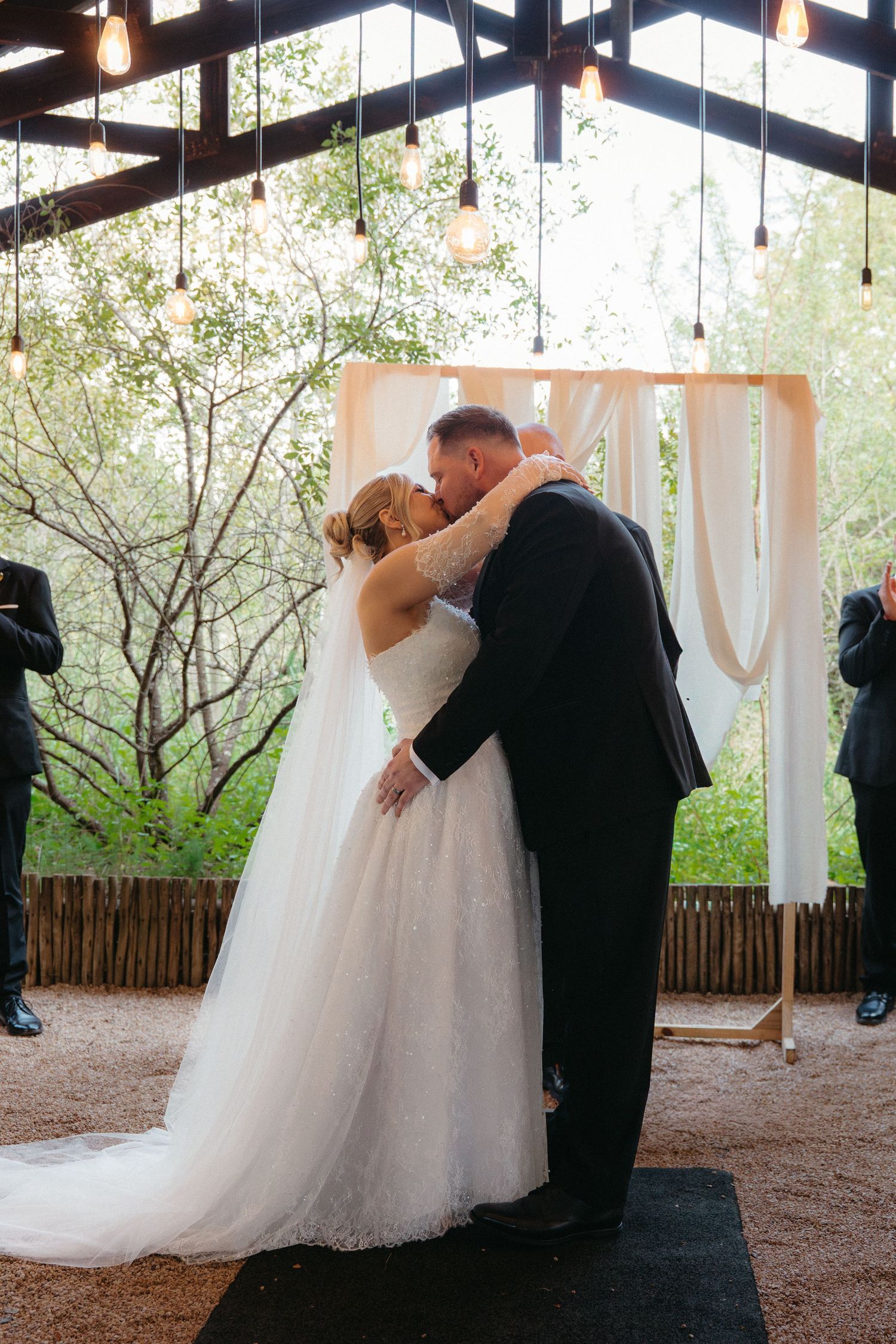 A couple shares their first dance under a rustic wooden pavilion surrounded by blooming trees.