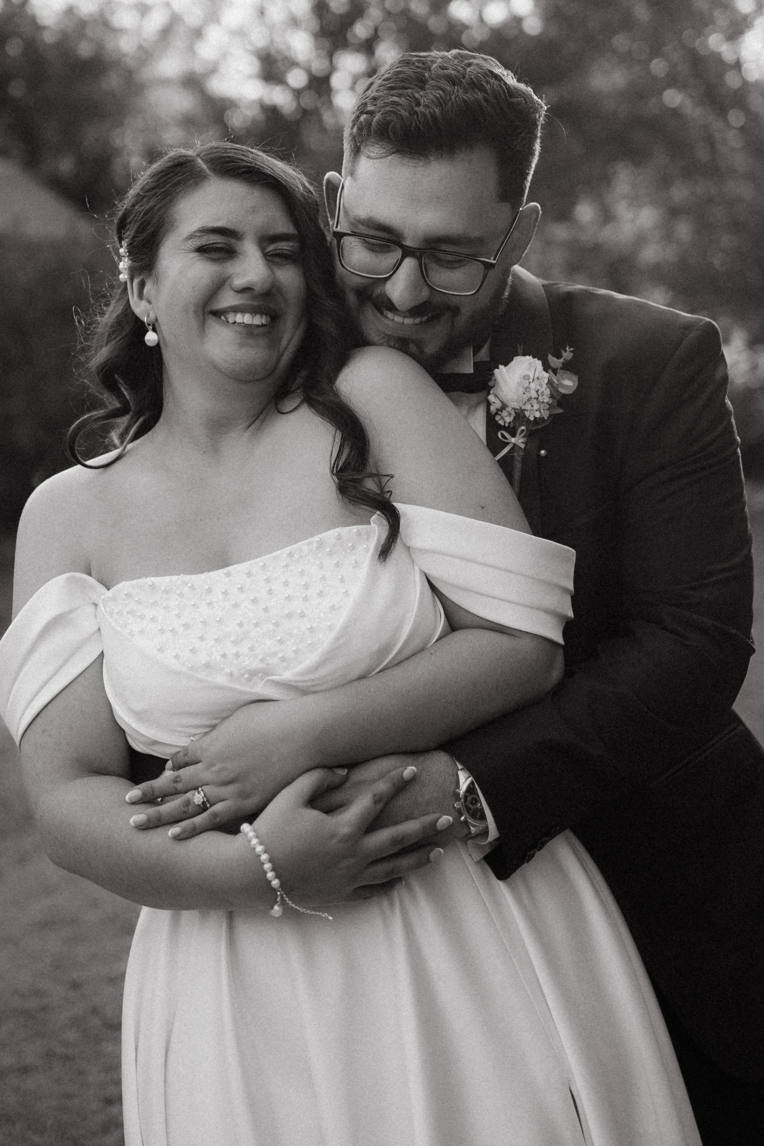 A tender black and white wedding portrait shows a couple embracing outdoors, the bride in an off-shoulder gown and groom in suit.