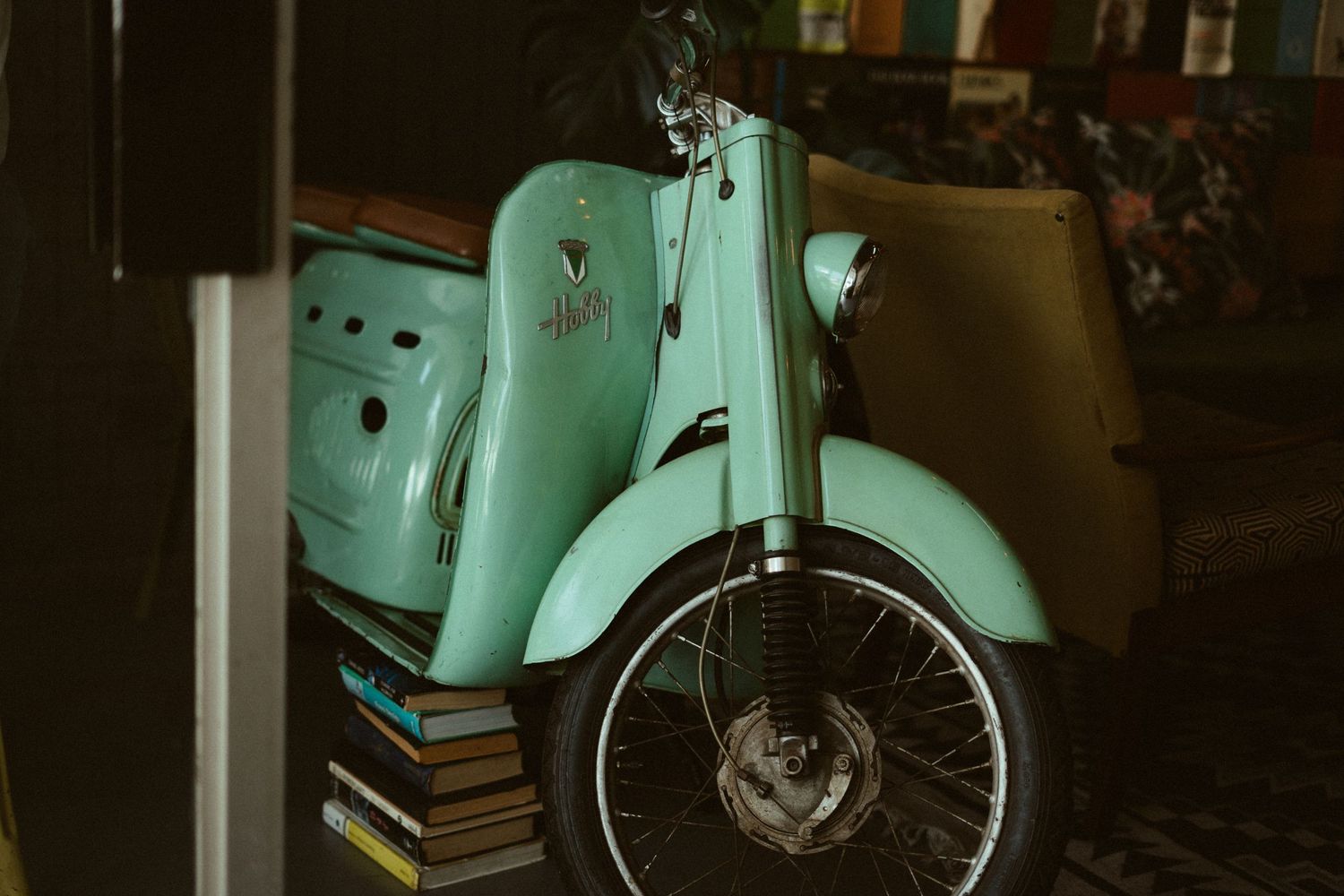 A vintage mint green Vespa scooter parked indoors next to stacked books with retro styling.
