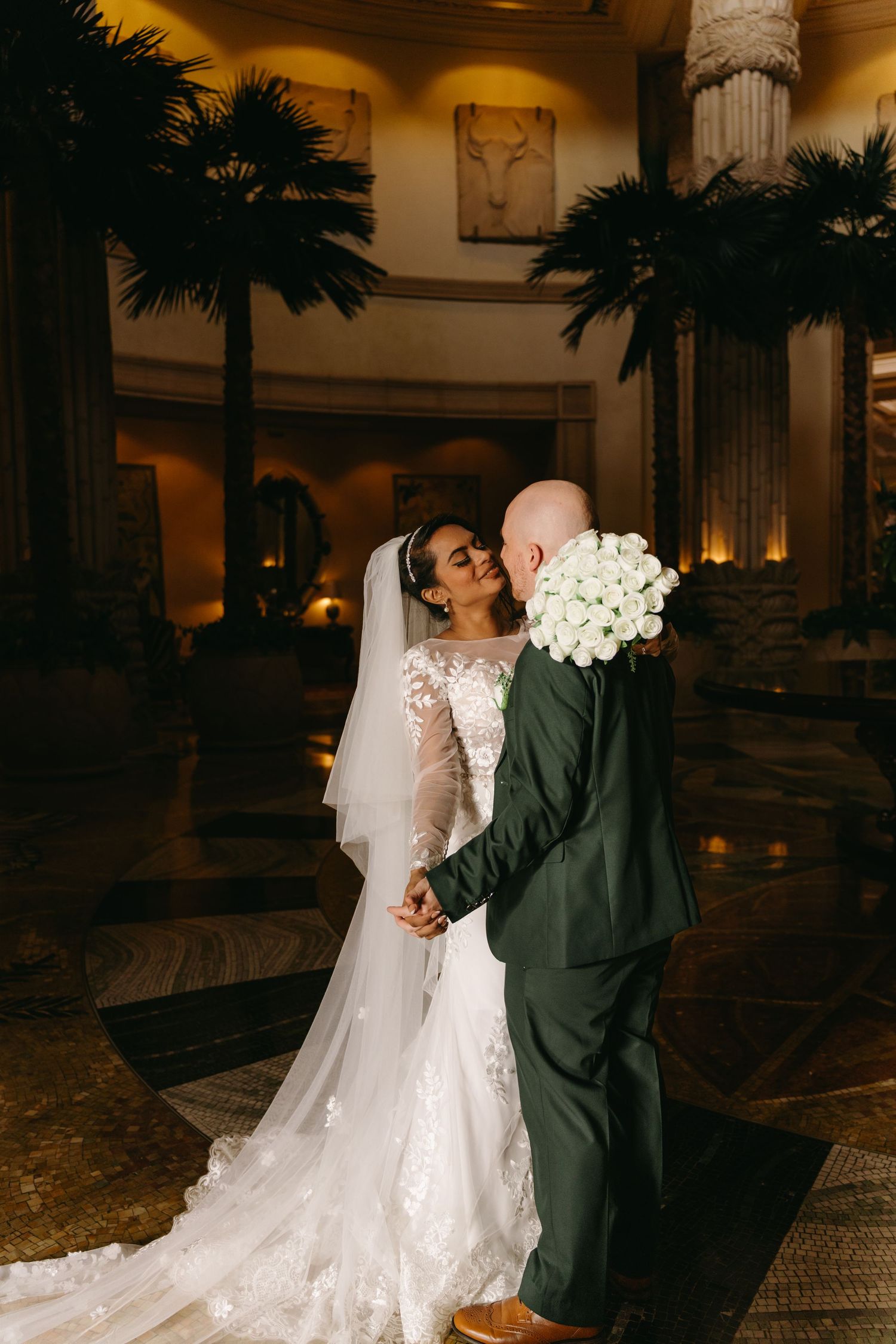 Bride and groom sharing romantic kiss in elegant hotel lobby at the Palace at Sun City with palm tree decor.