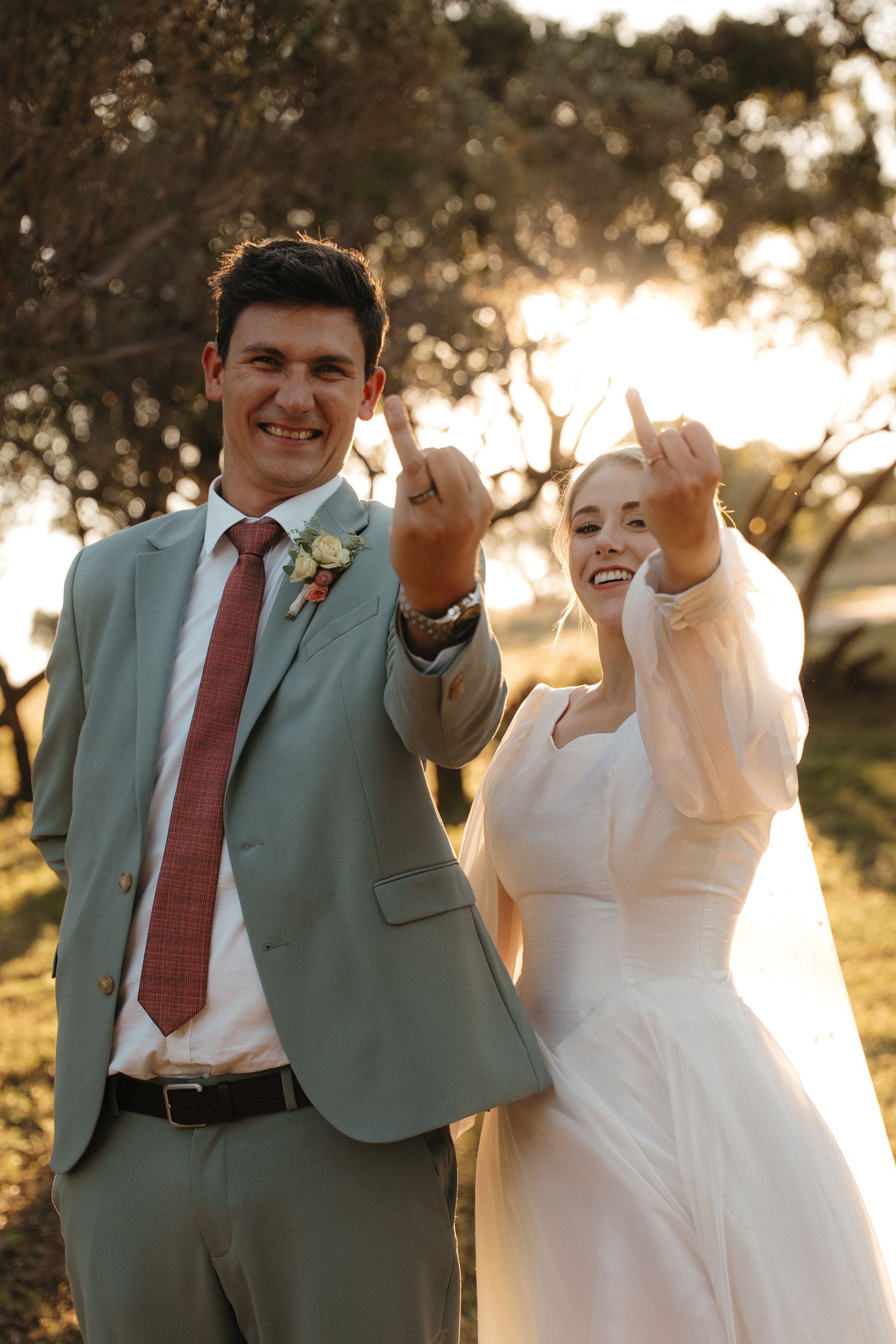 Smiling bride and groom in wedding attire playfully holding uo ring fingers outdoors at golden hour in Rustenburg