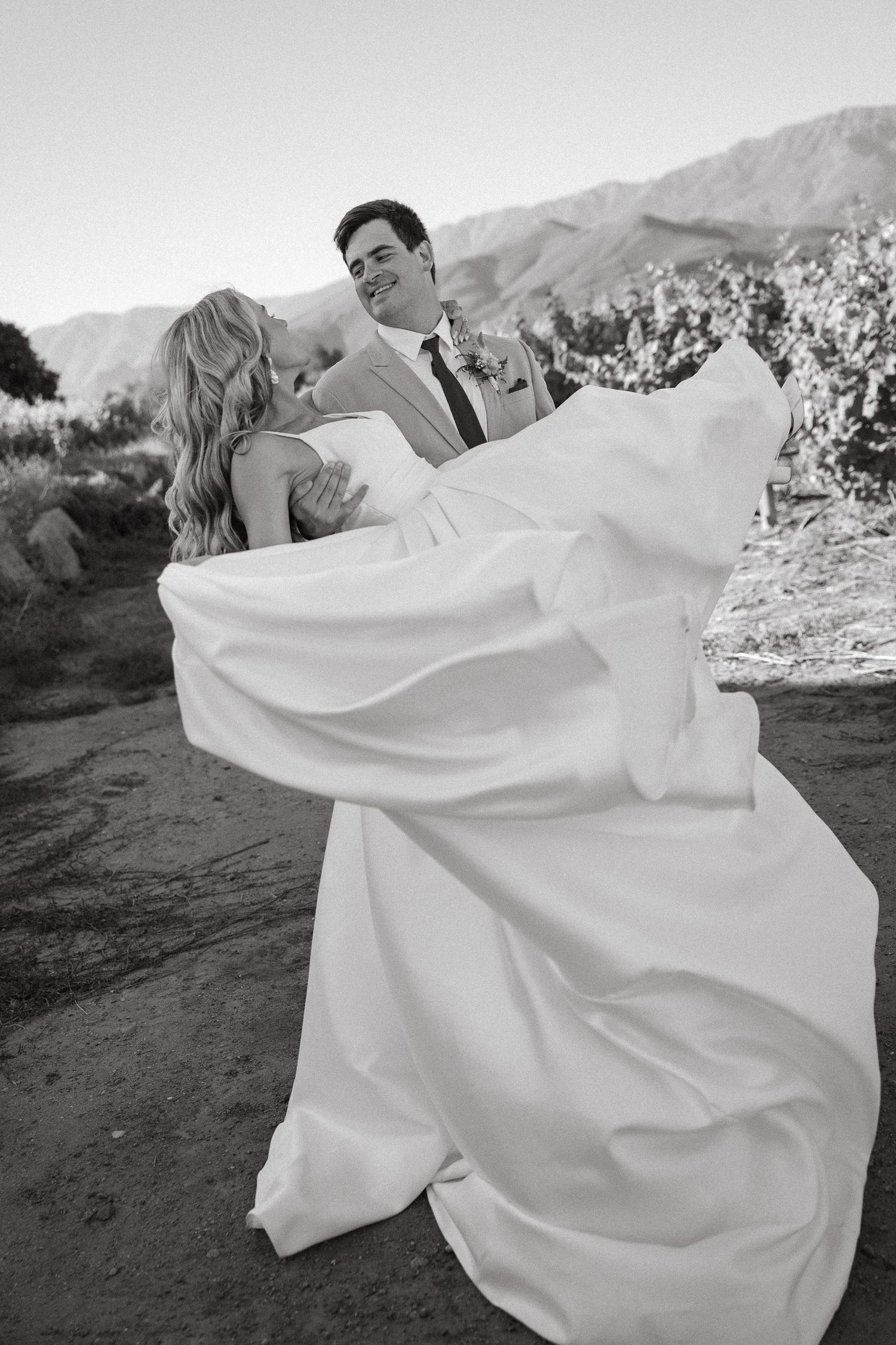 Black and white photo of a groom in a suit twirling his bride in a flowing satin gown outdoors, with vineyards and mountains behind them.