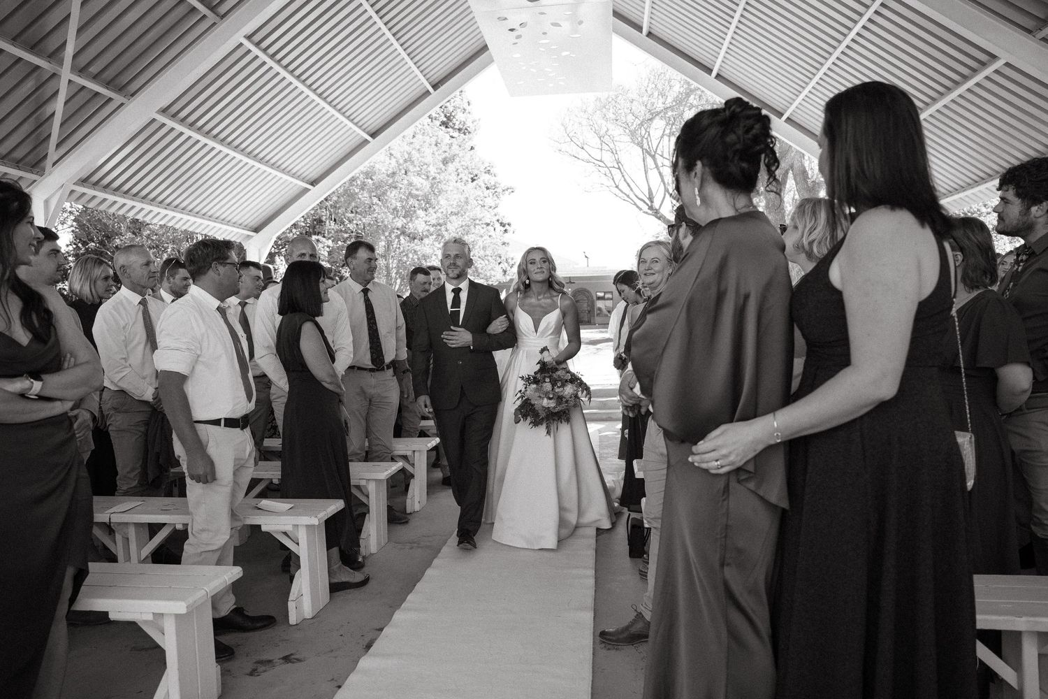 Father walks bride in white gown with bouquet down the aisle of an open-air pavilion as wedding guests stand beside wooden benches.