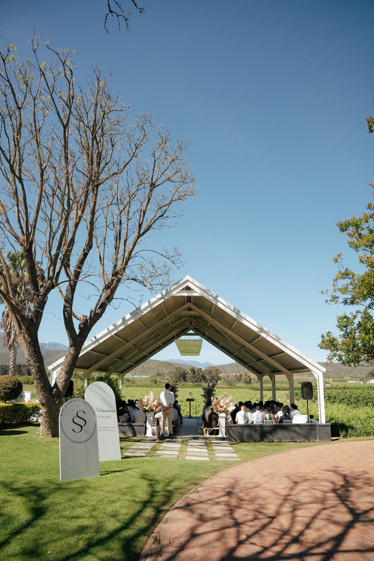 Outdoor wedding ceremony under an open-sided pavilion with guests seated, beside a bare tree and monogram signs on a lawn near vineyards.