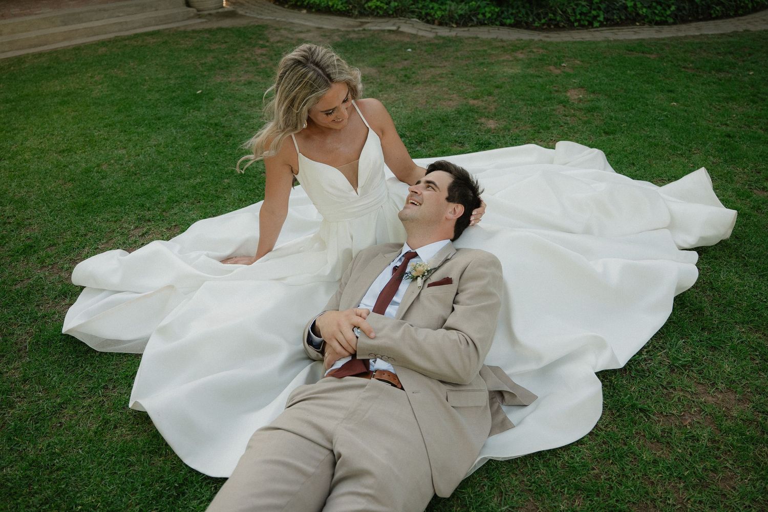 Bride in white gown sits on grass beside groom in tan suit lying down, sharing a romantic moment outdoors.
