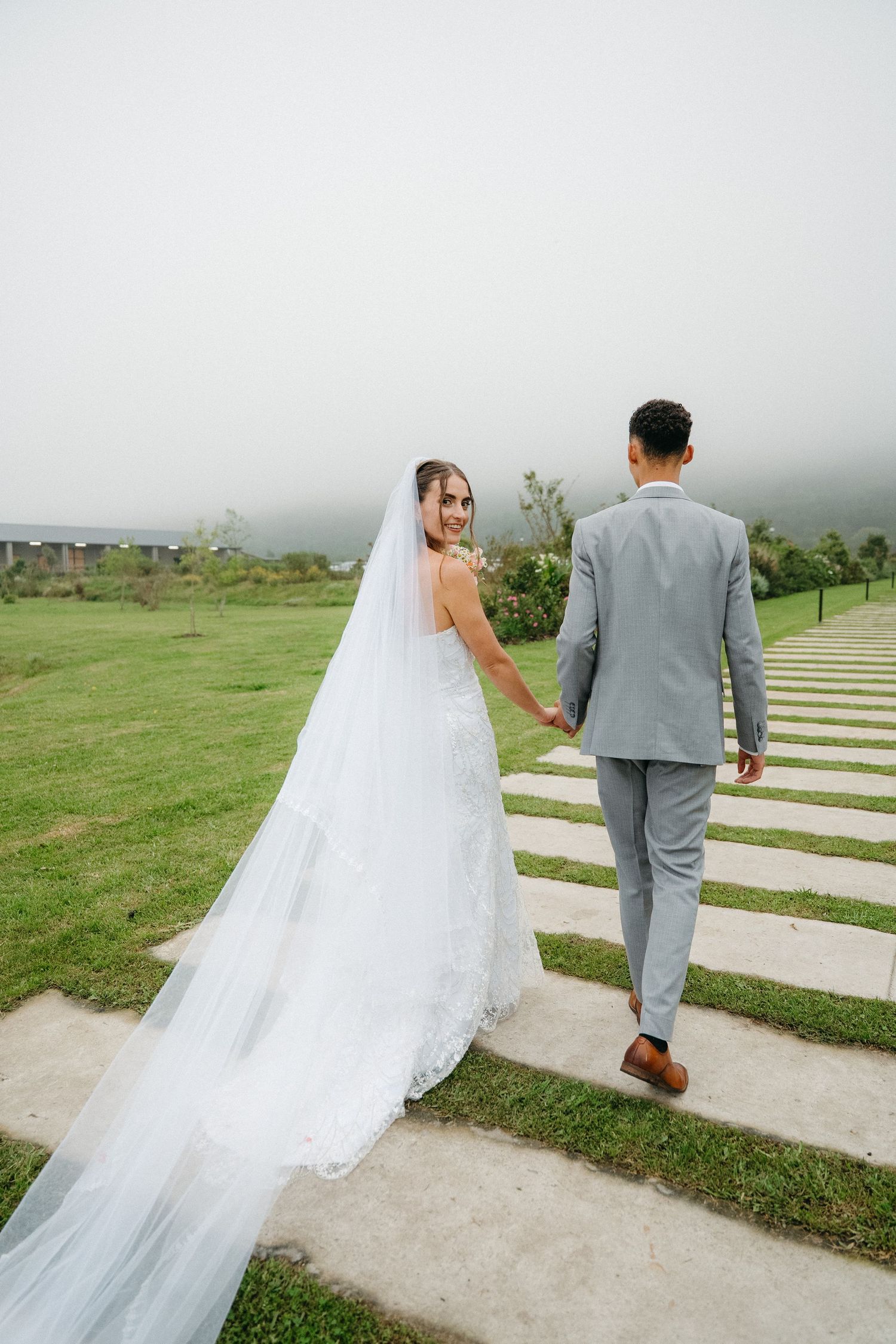 Bride in white gown and veil holds grooms hand walking up stone path on lush green lawn on misty day.