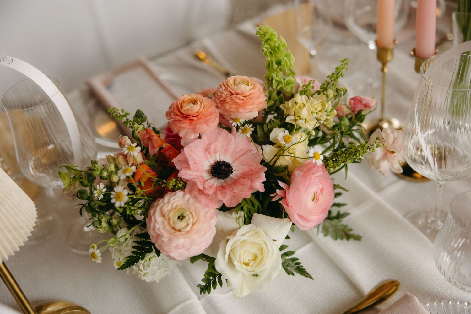 Pink ranunculus and anemone wedding centerpiece with white roses and greenery on an elegant table setting.