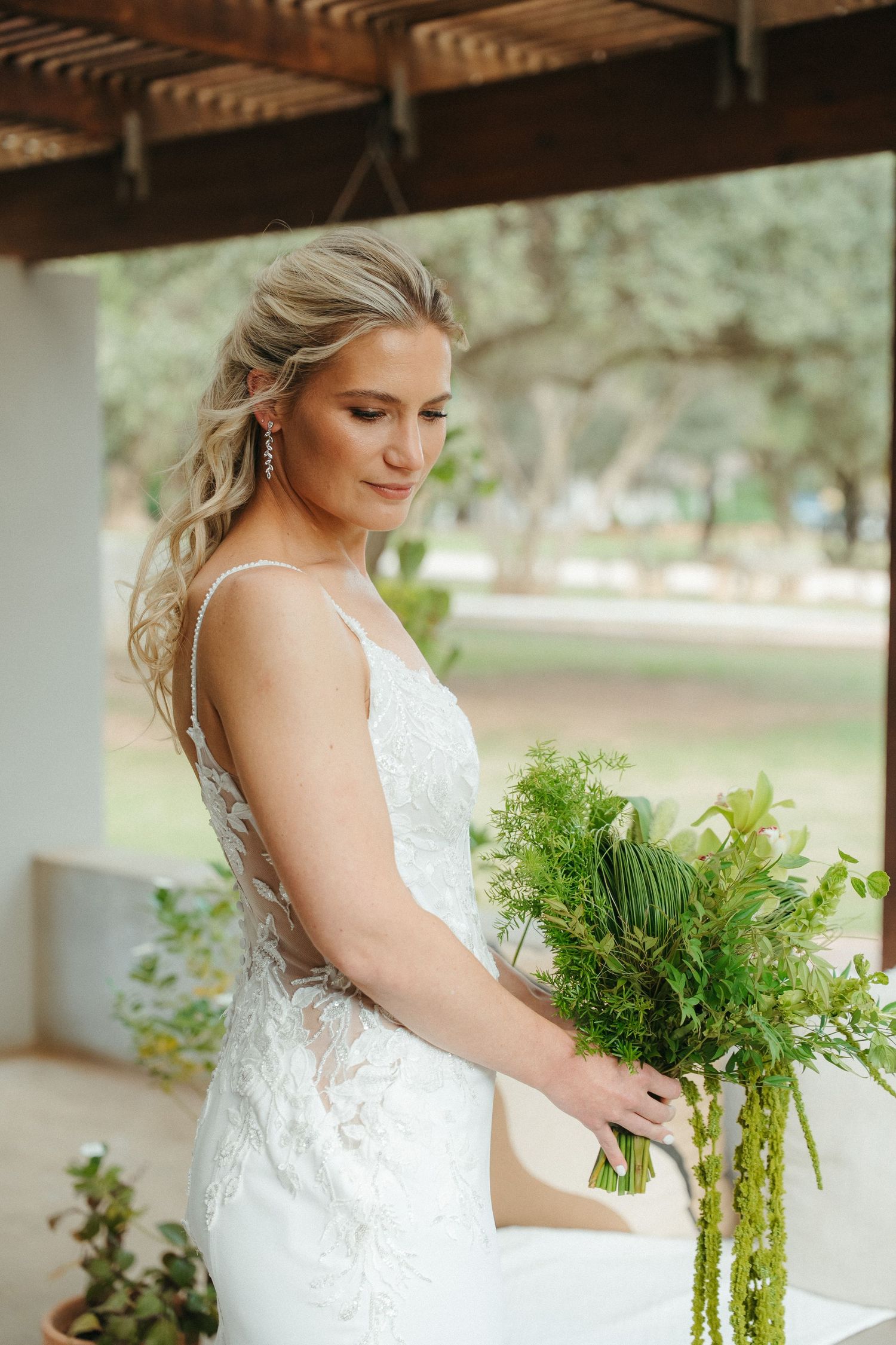 Bride in white lace gown holding green bouquet under wooden pergola.