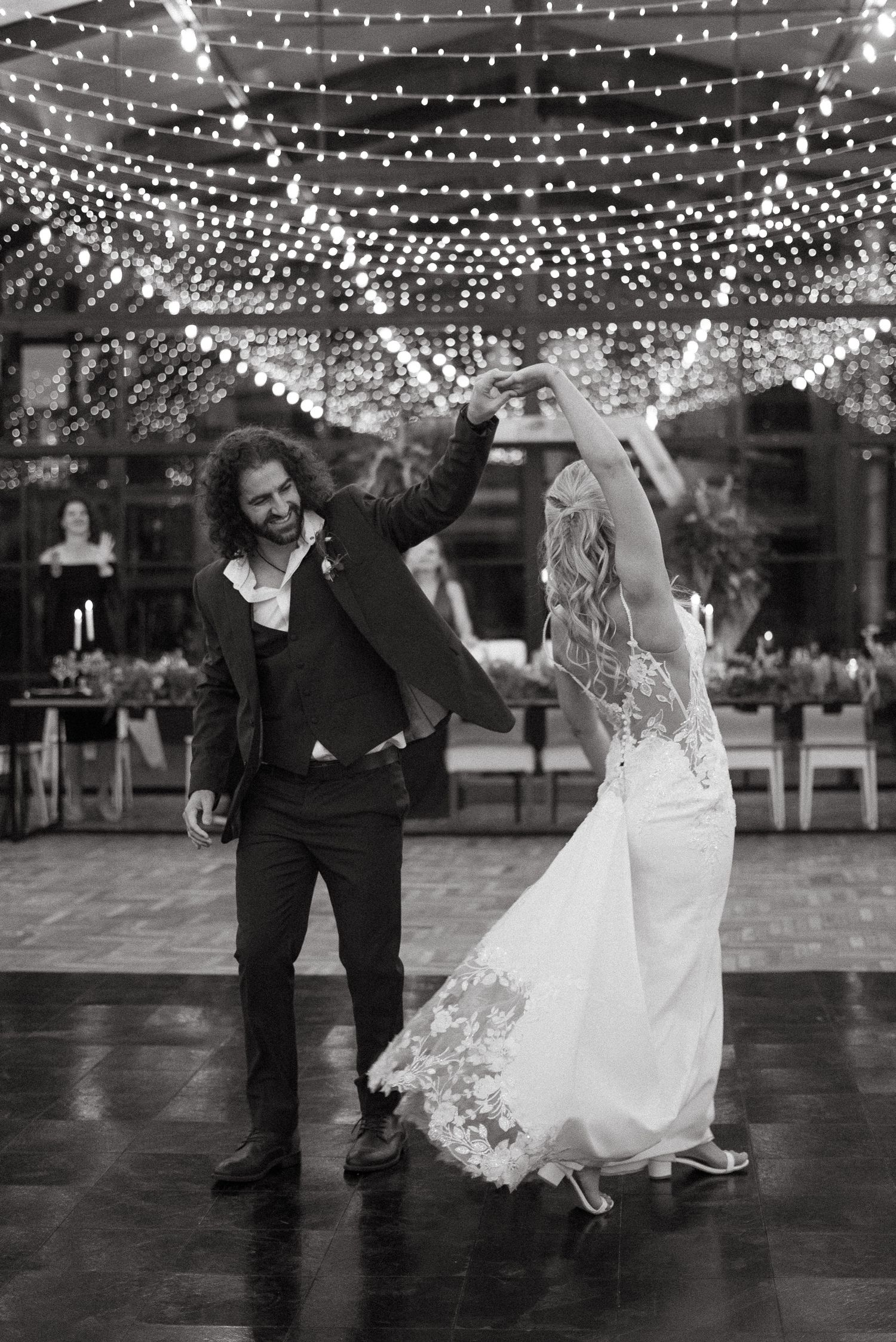 Black and white photo of bride and groom sharing first dance under twinkling string lights at elegant wedding reception.