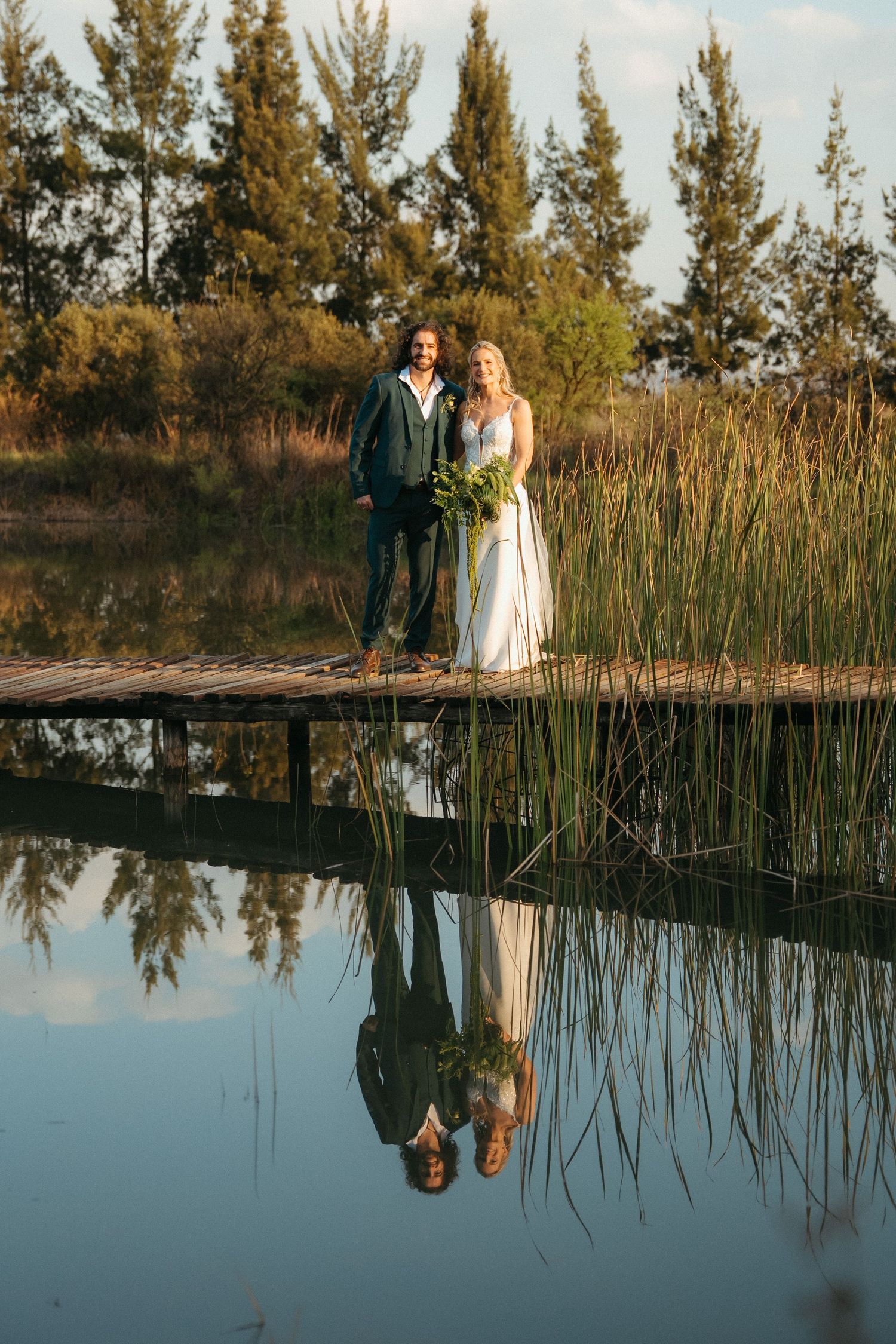 Bride and groom standing on a wooden dock reflected in calm water, surrounded by tall reeds and pine trees.
