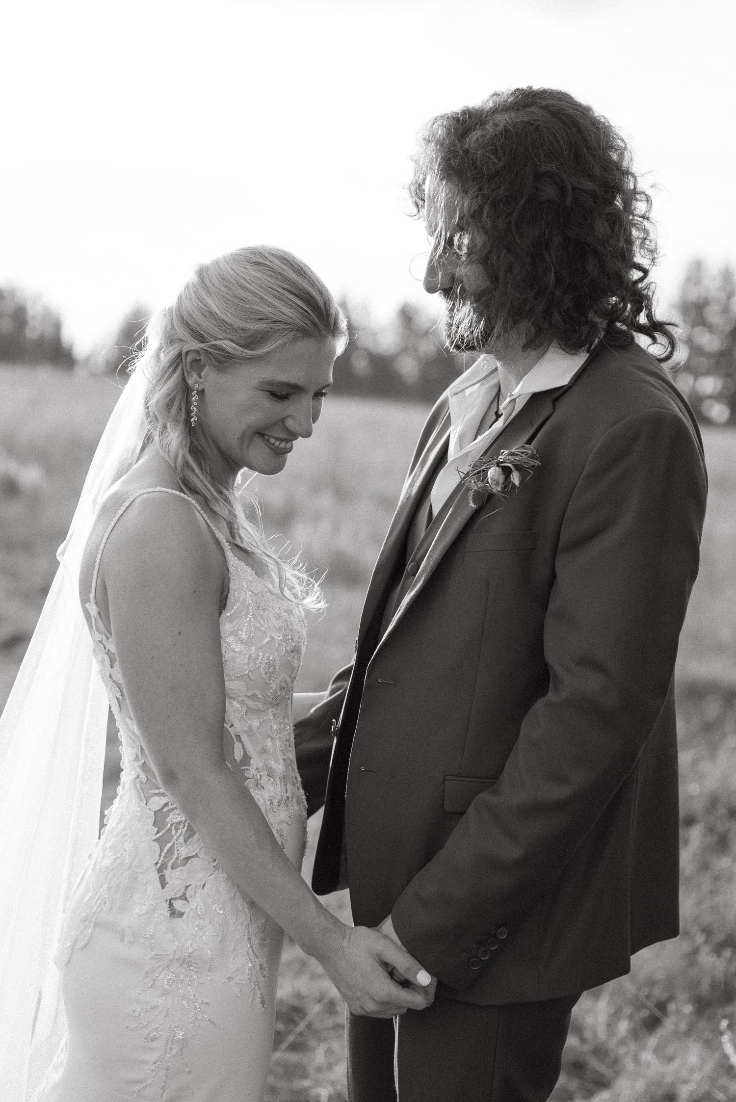 Black and white photo of a smiling bride in lace gown and groom with curly hair holding hands outdoors.