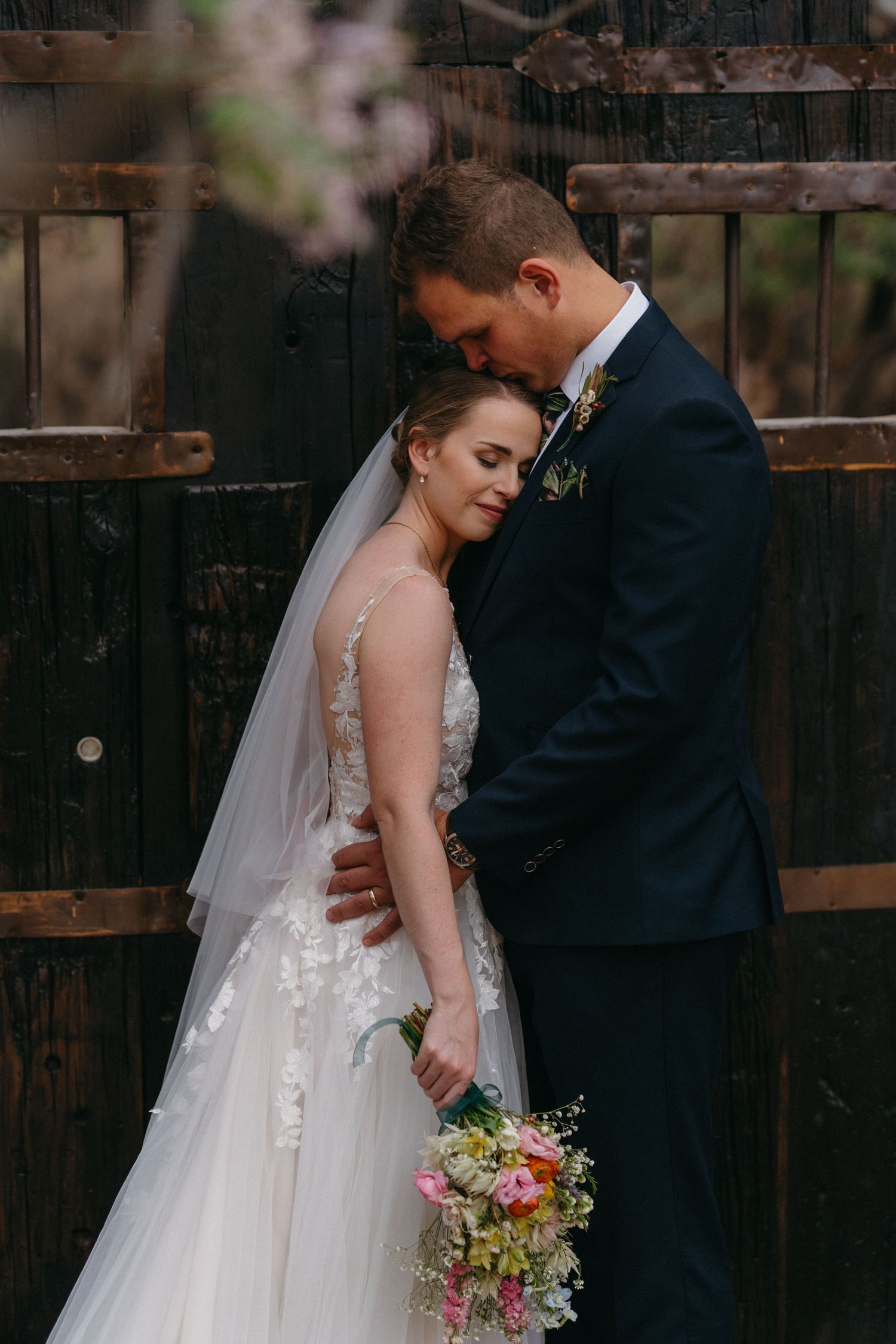 Romantic bride and groom embracing tenderly against rustic wooden fence with spring blossoms in Dinokeng, Pretoria.