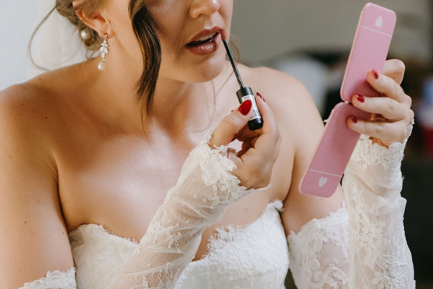Someone in a white lace wedding dress applies lipstick while holding a pink compact mirror.