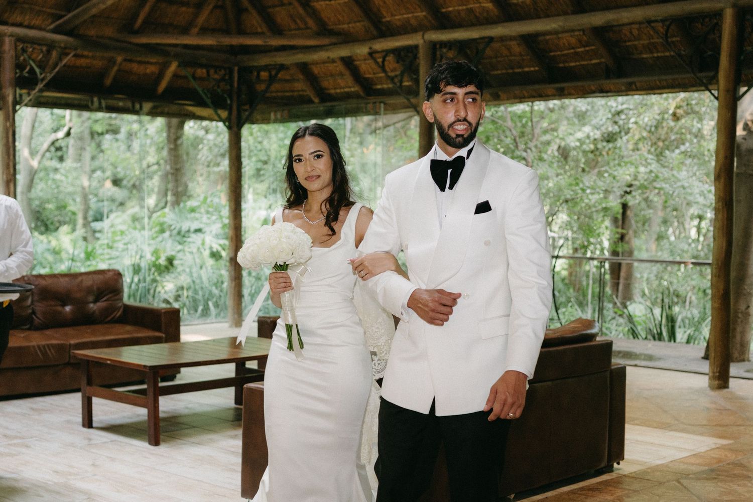 Couple in white wedding attire poses under rustic wooden gazebo at outdoor venue.