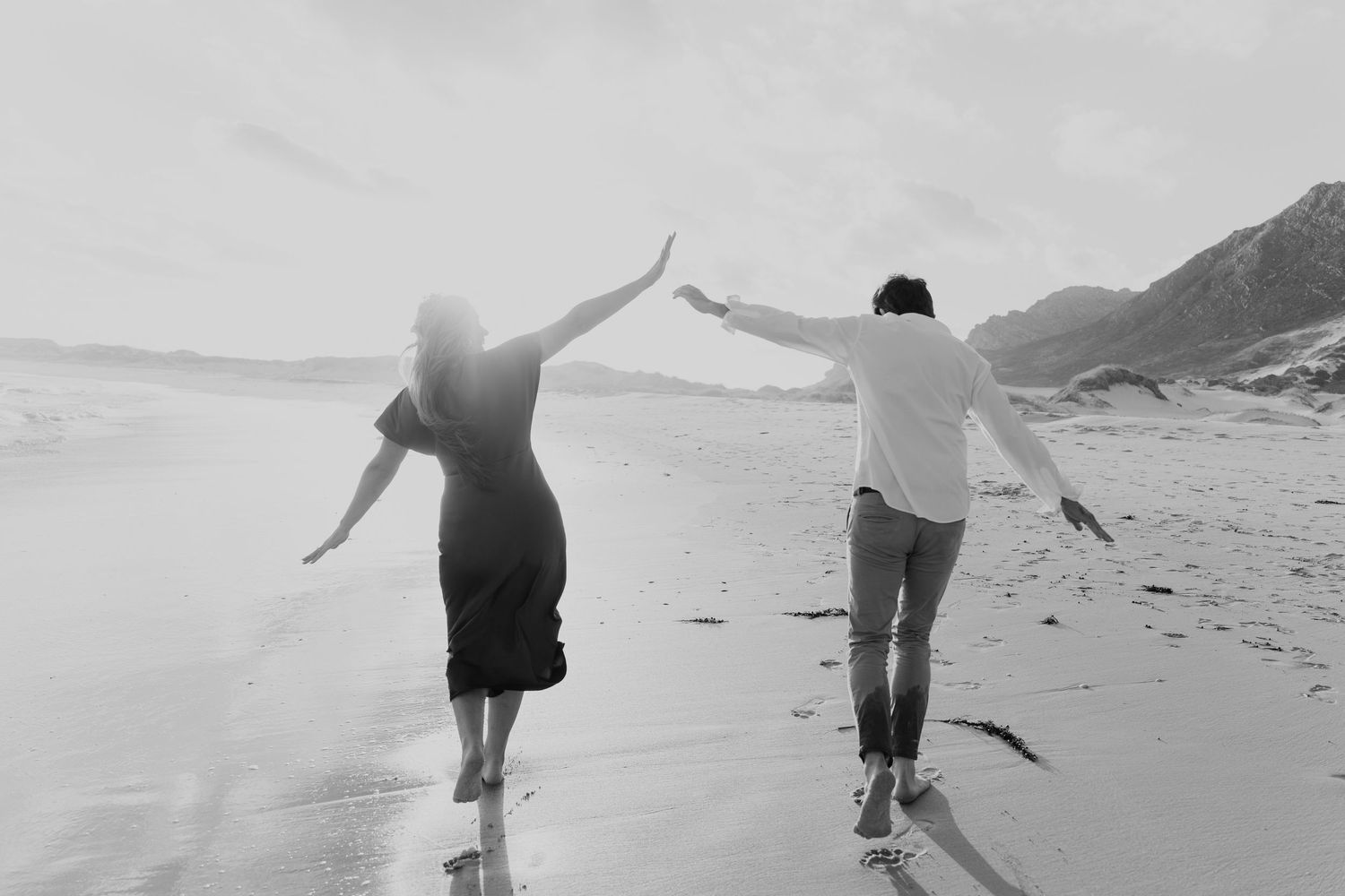Black and white photo of a couple holding hands and walking barefoot on a beach with mountains in the background.