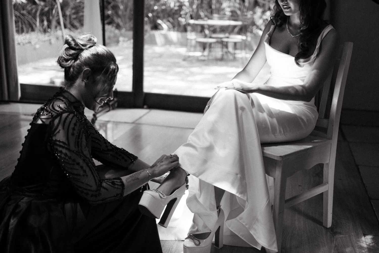 A black and white photograph shows someone in a dress adjusting another person's footwear in a sunlit room.