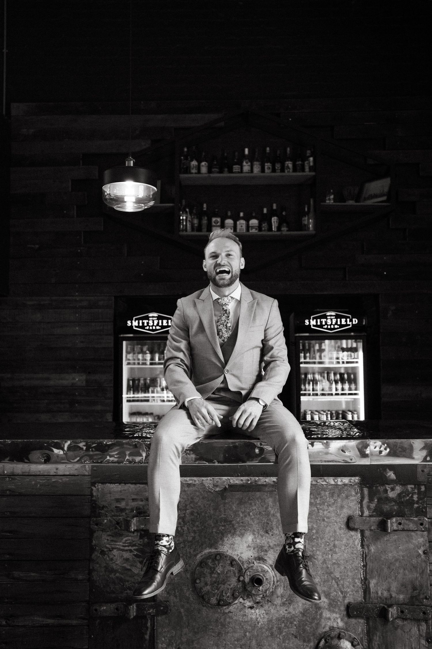 A groom in a light gray suit sits casually in front of illuminated bar shelves.
