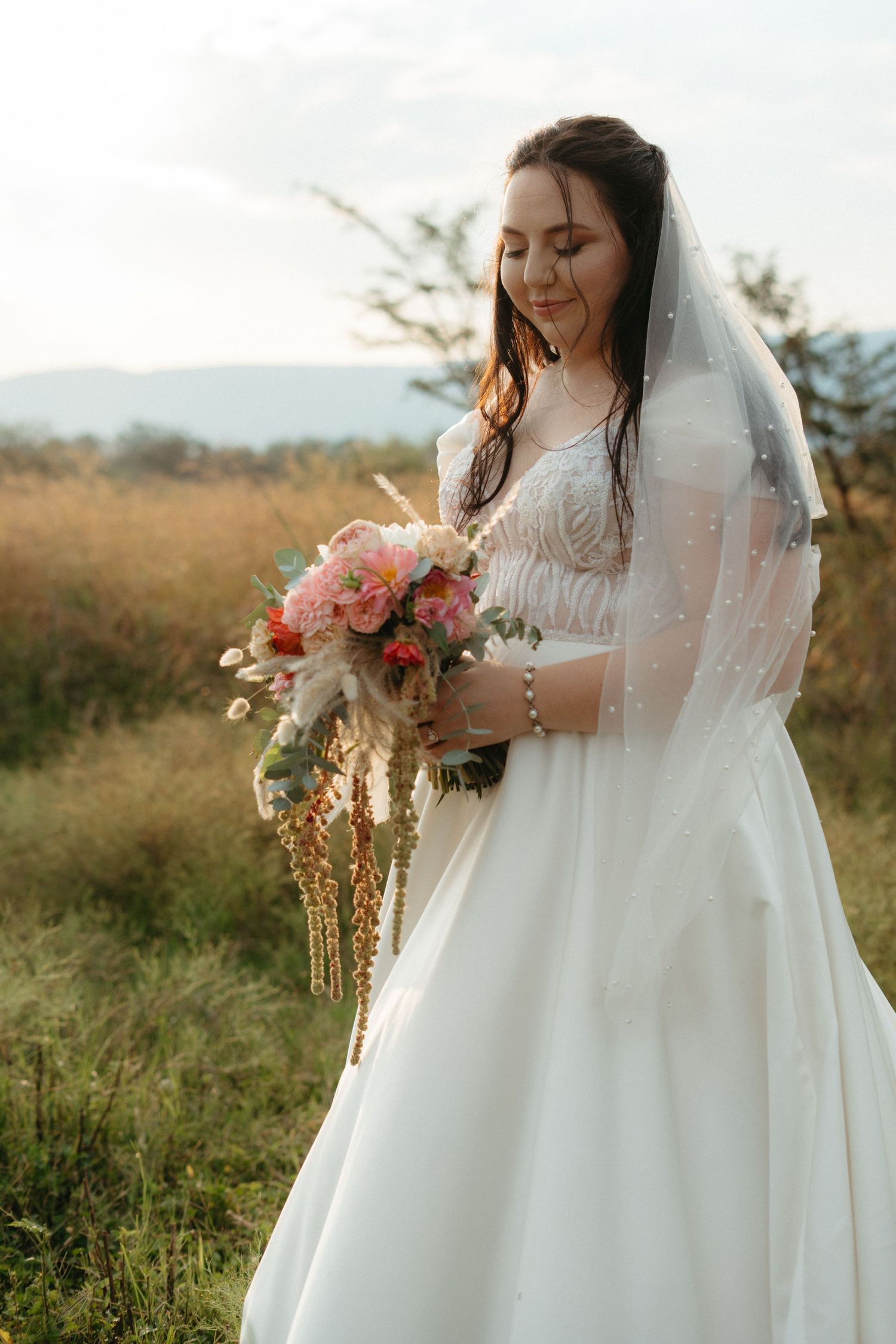 A wedding dress and veil silhouetted against mountains with a bouquet of pink and peach flowers at golden hour.