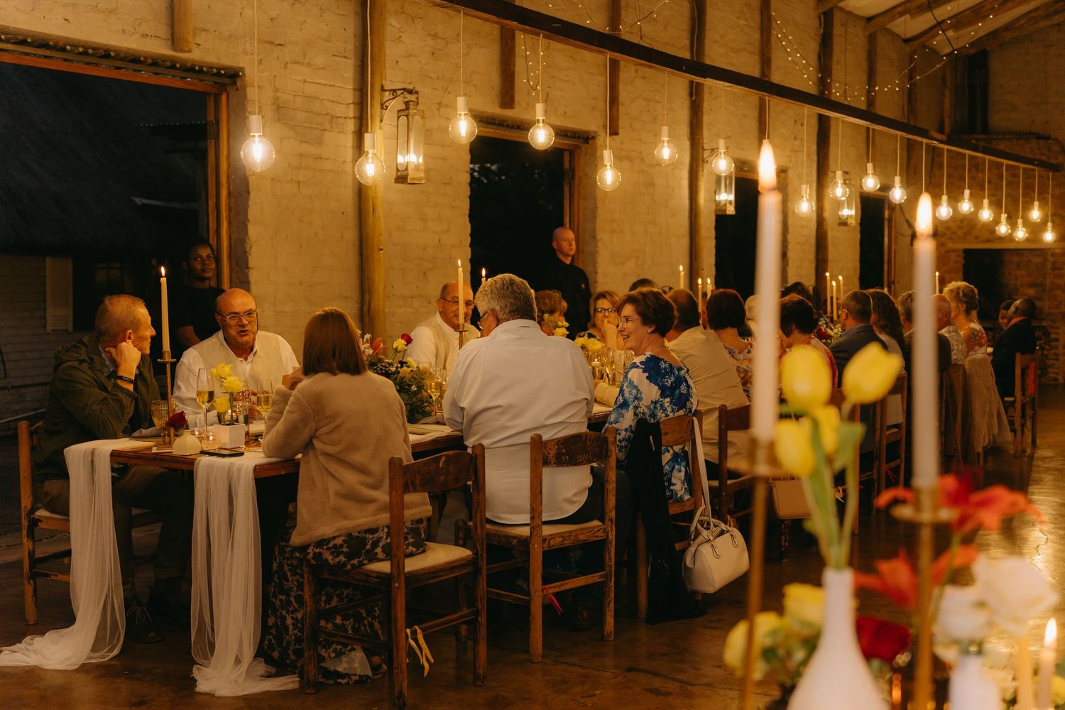 Rustic barn wedding reception with hanging lights and guests dining at long tables.