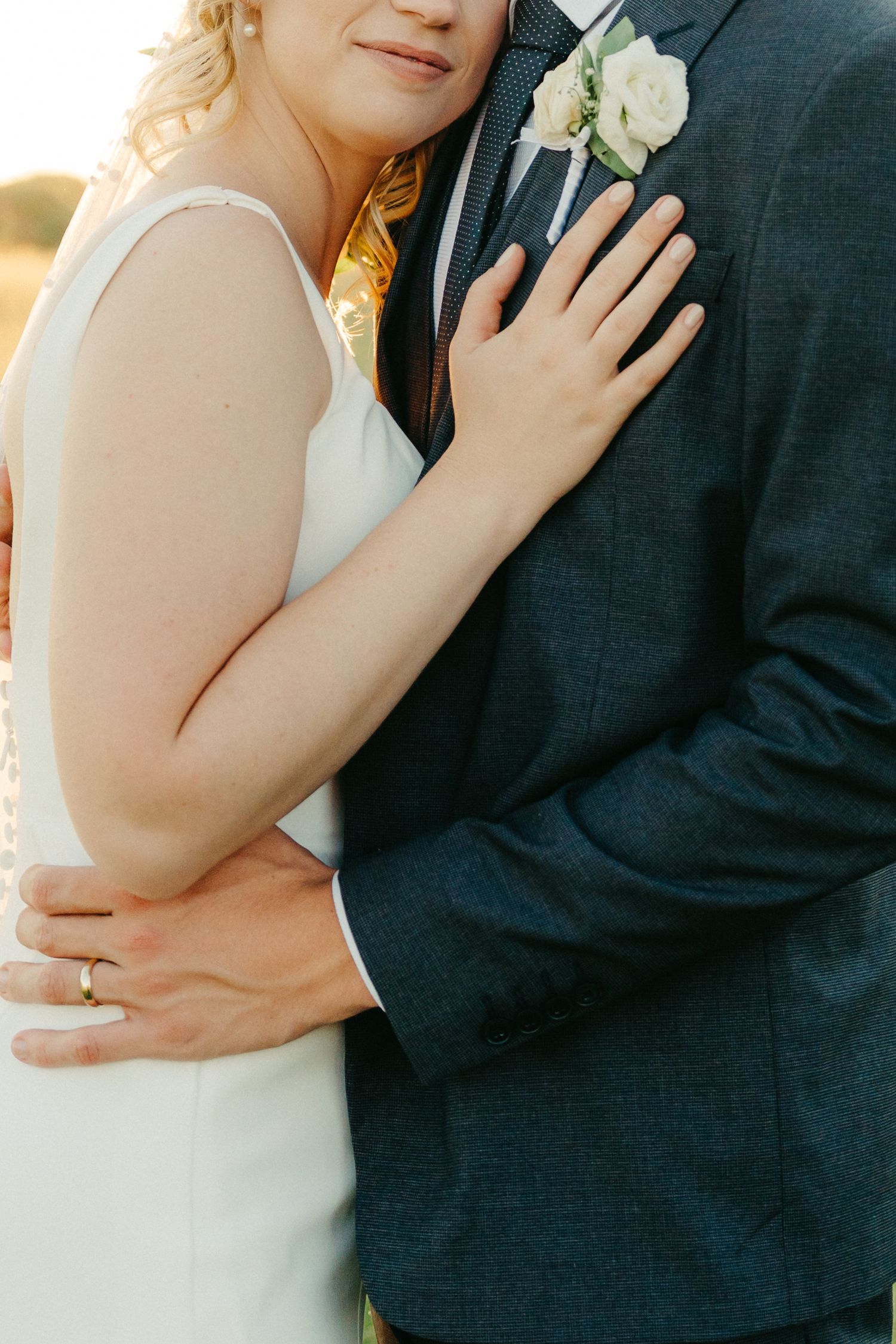 Close up of a couple embracing, showing the bride's hand on the groom's boutonniere and navy suit.