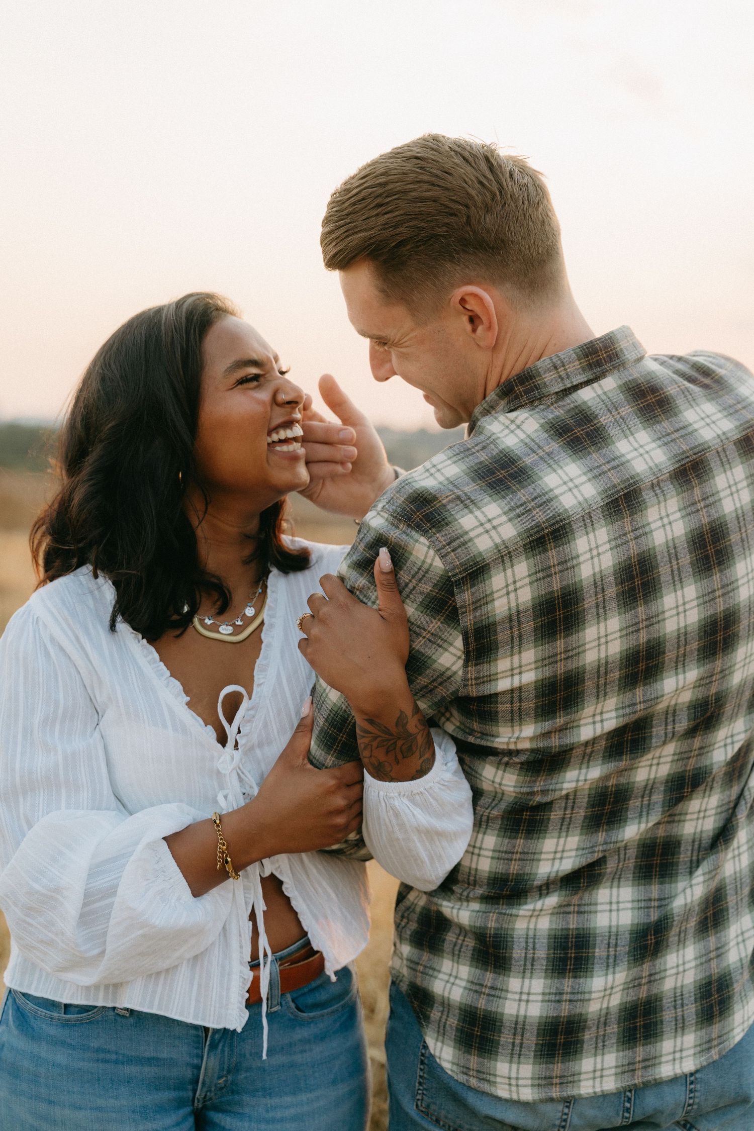 A couple shares playful intimate moments in a field during sunset while wearing casual attire.