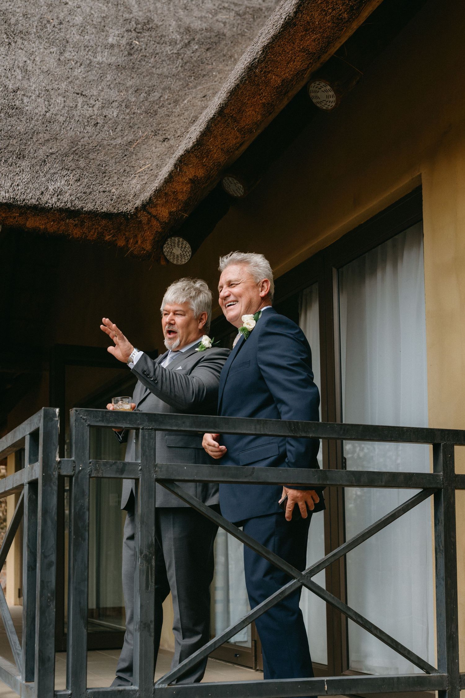 Men in formal suits gesture enthusiastically from a balcony with thatched roof.