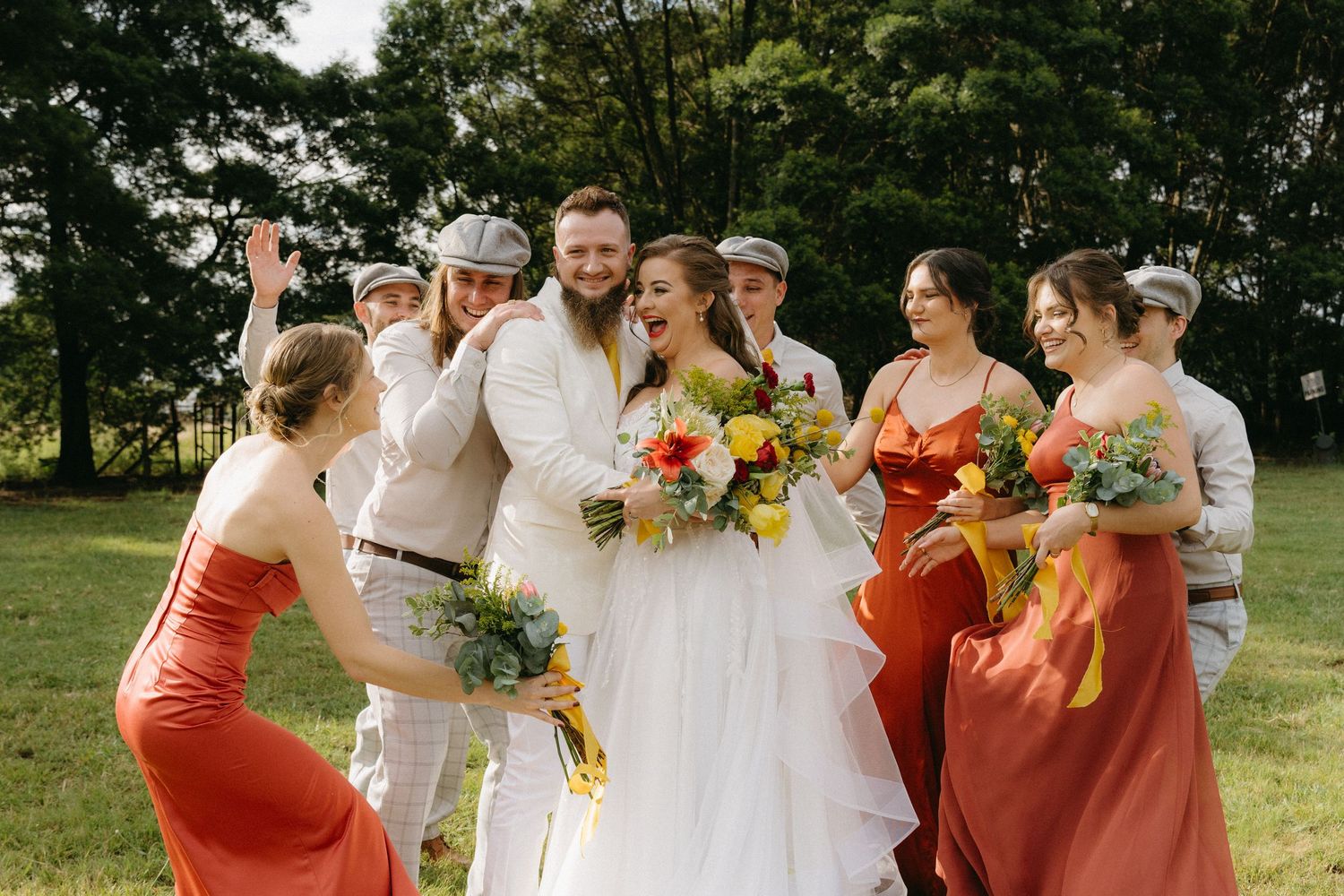 Wedding party poses on sunny lawn with bridesmaids in coral dresses holding vibrant summer bouquets.