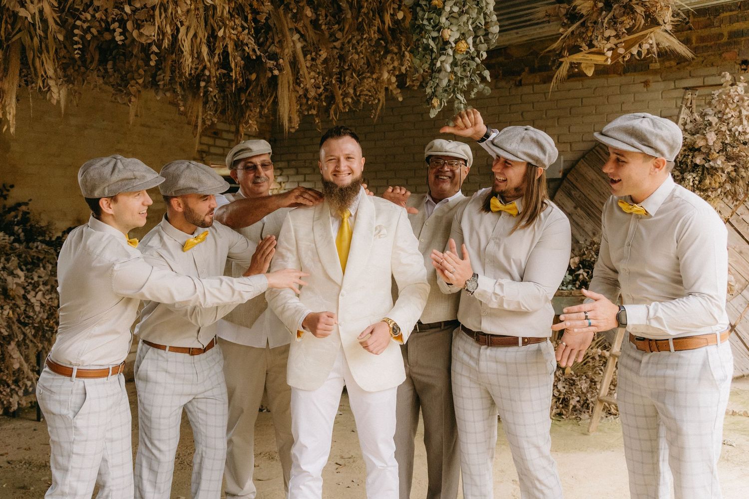 Groomsmen in white outfits and newsboy caps toasting and celebrating at rustic wedding venue.