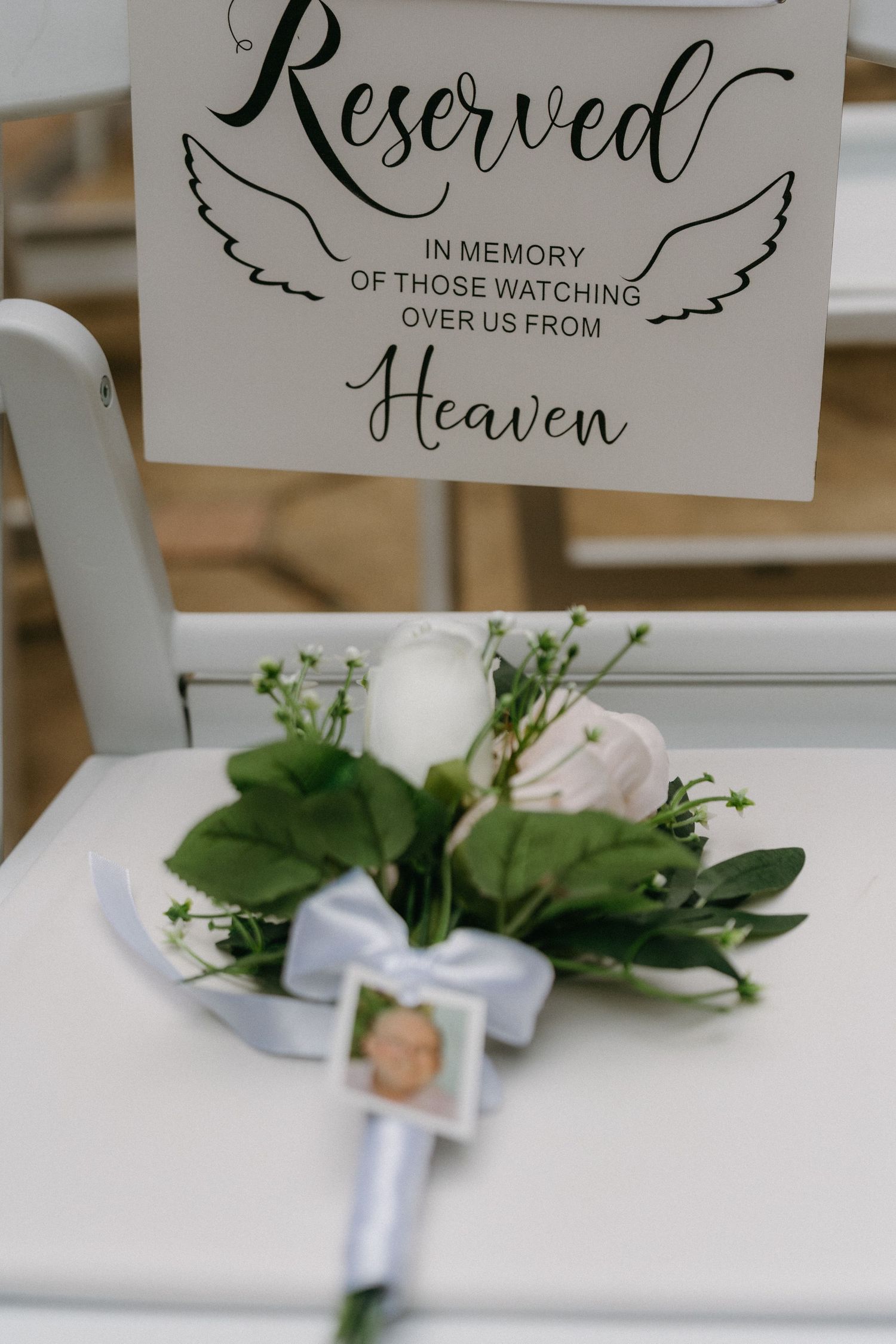 Reserved chair displays touching memorial signage with greenery and white bow accents at wedding ceremony setting.