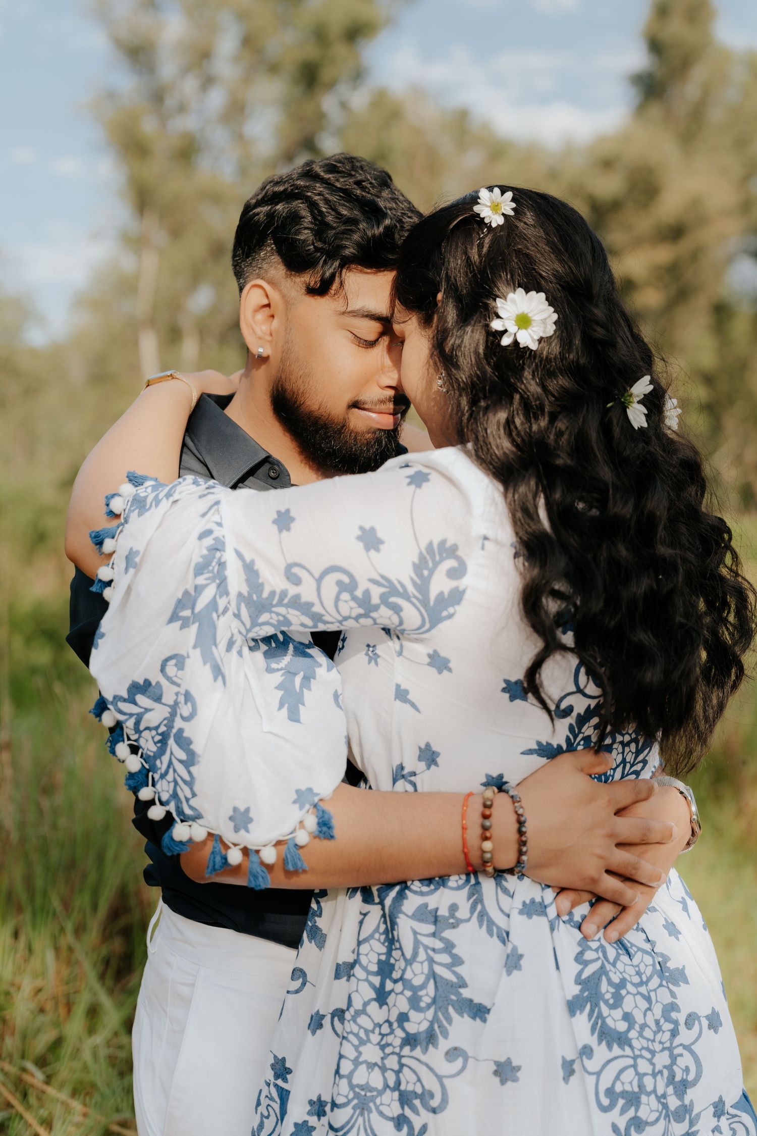 A romantic couple embraces in a field, with the woman wearing a blue and white floral dress and daisies in her dark hair.