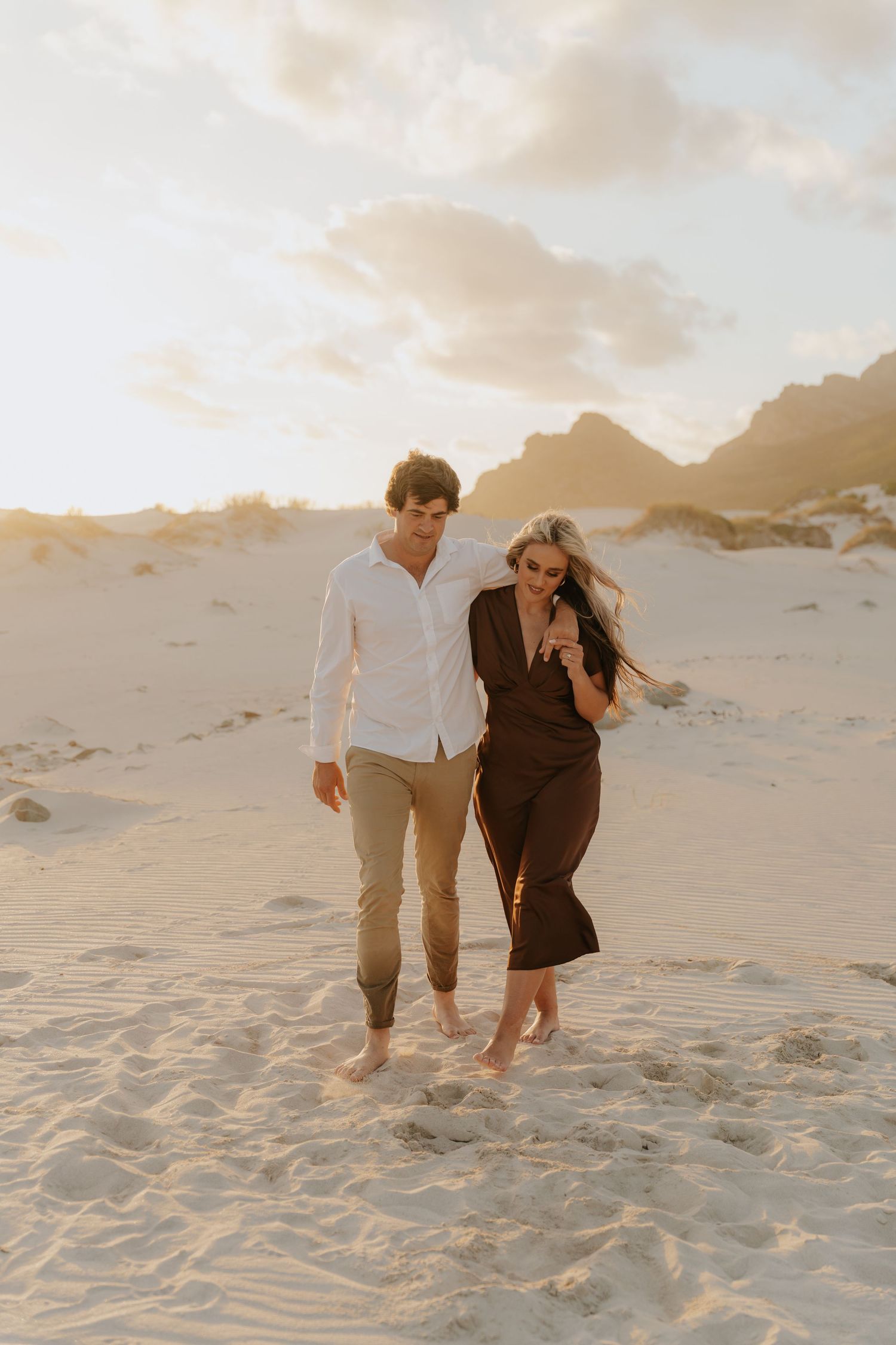 Couple walking barefoot on sandy beach at golden sunset, man in white shirt and woman in dark jumpsuit.