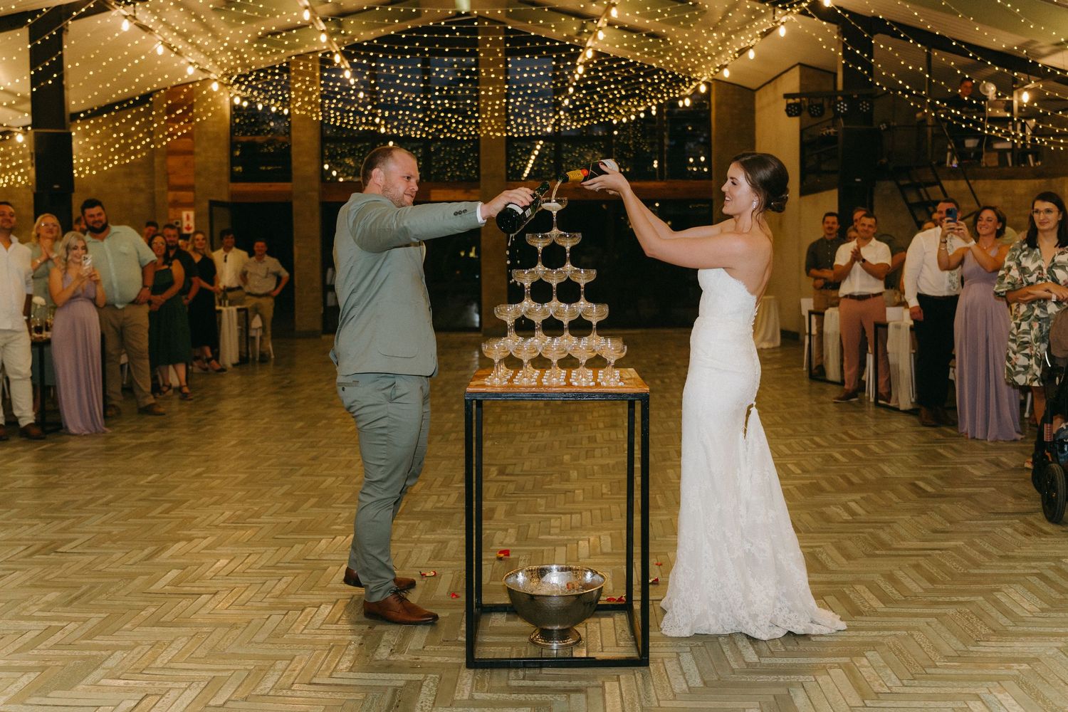 Wedding celebration with couple pouring champagne into tower of glasses at reception venue with fairy lights overhead.