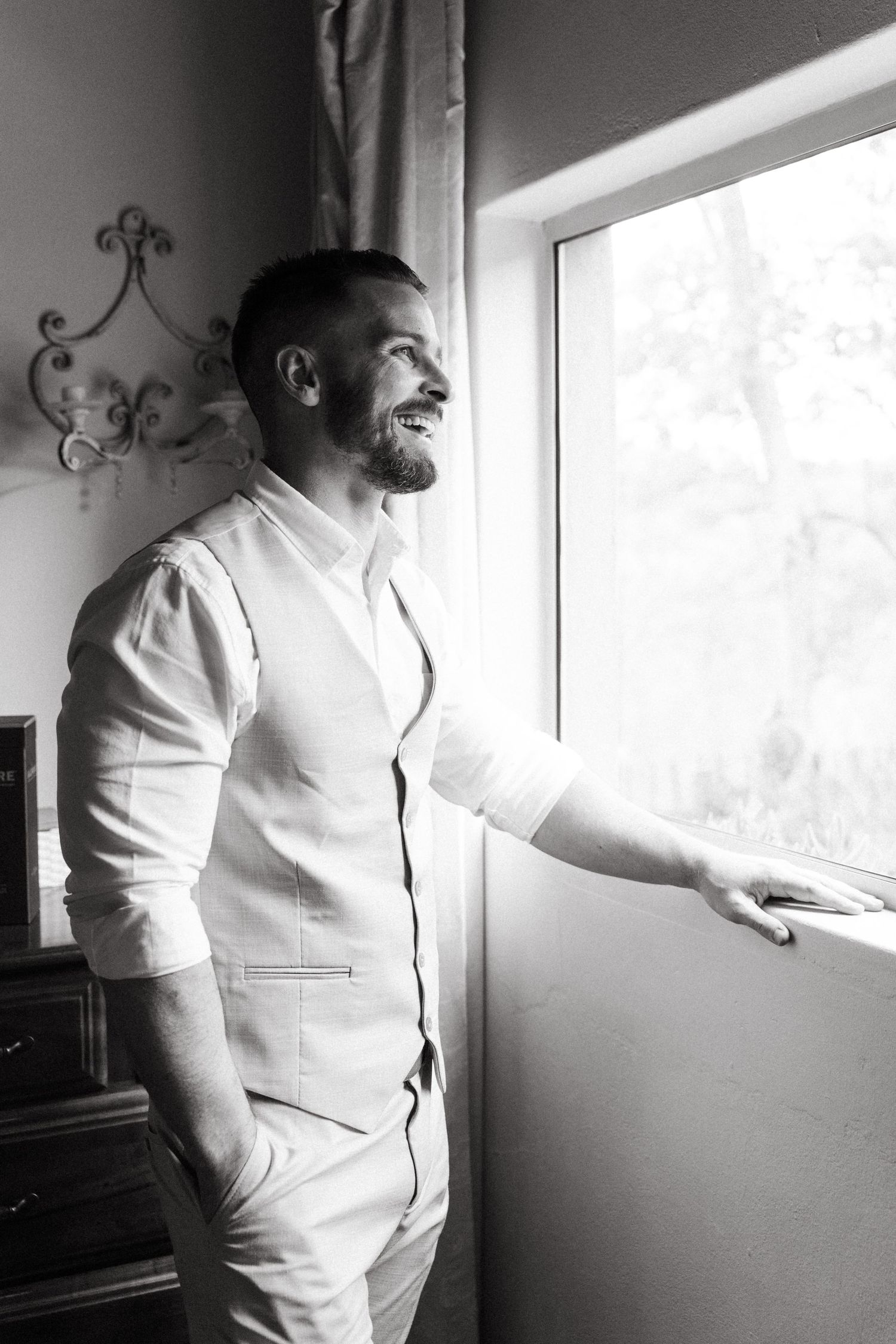 Groomsmen in light colored suits look out windows in an elegant getting-ready room.