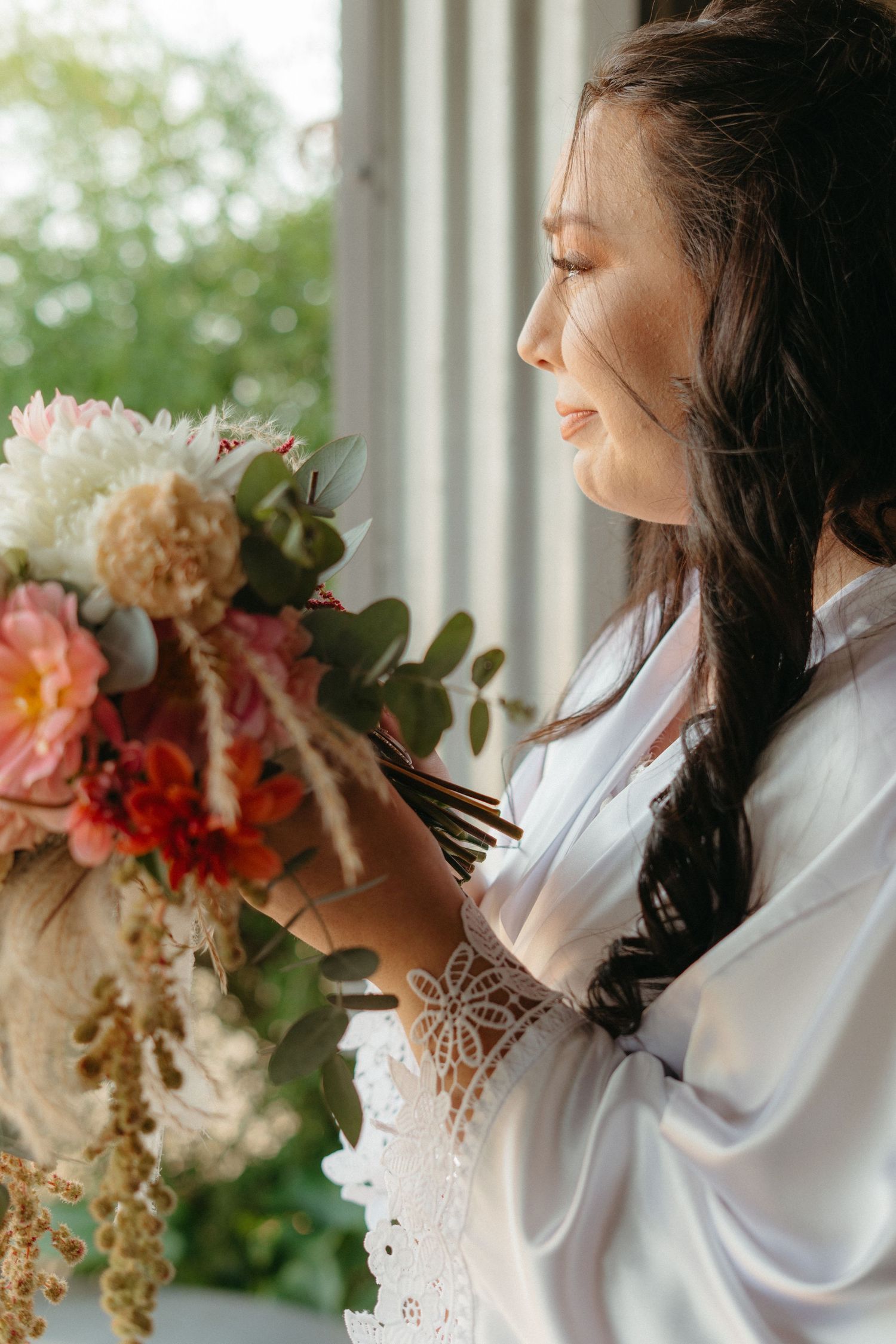 A joyful bride in a white robe holds a beautiful bouquet of mixed flowers by a window.