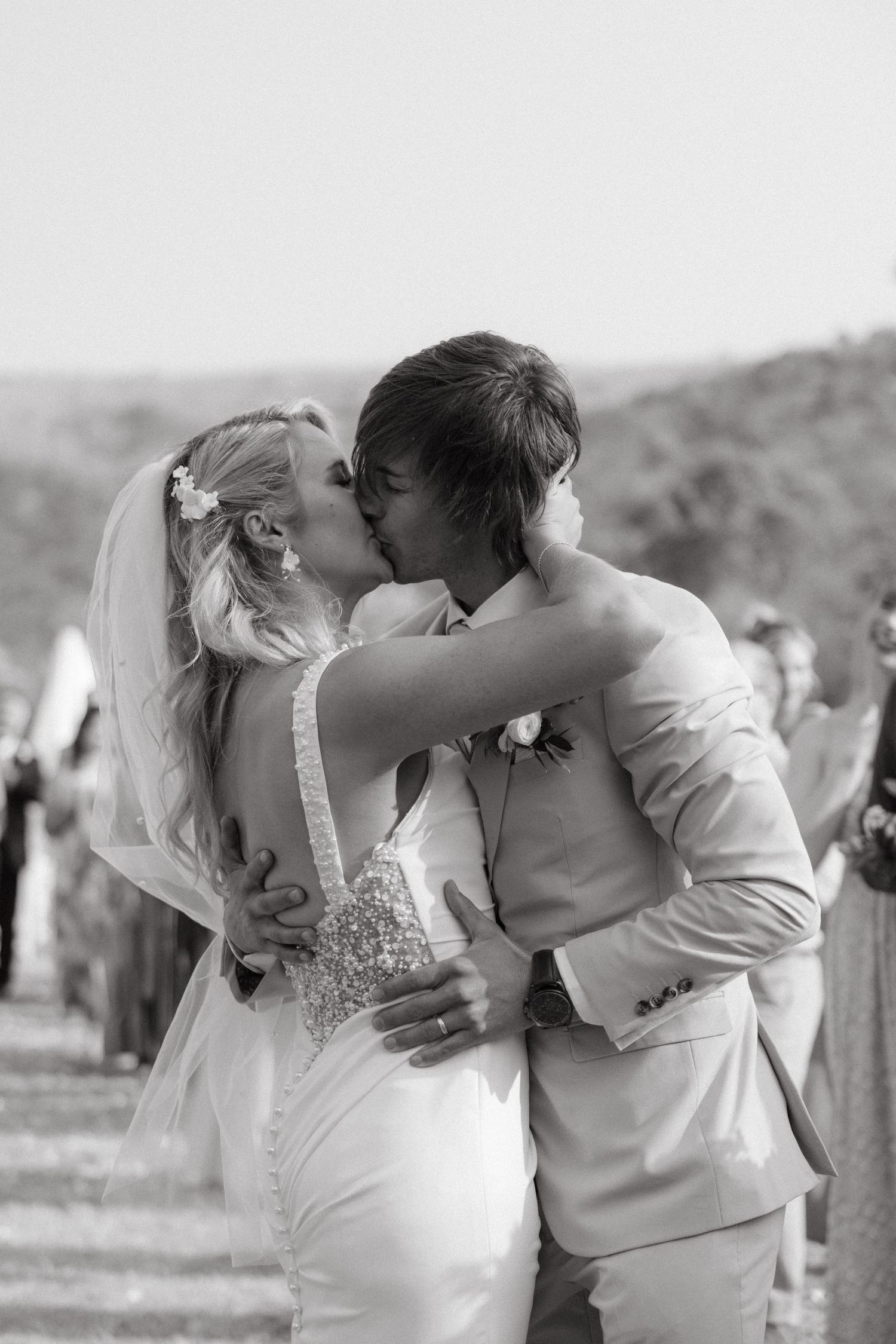 A romantic black and white wedding photo of a couple sharing their first kiss in an outdoor setting.
