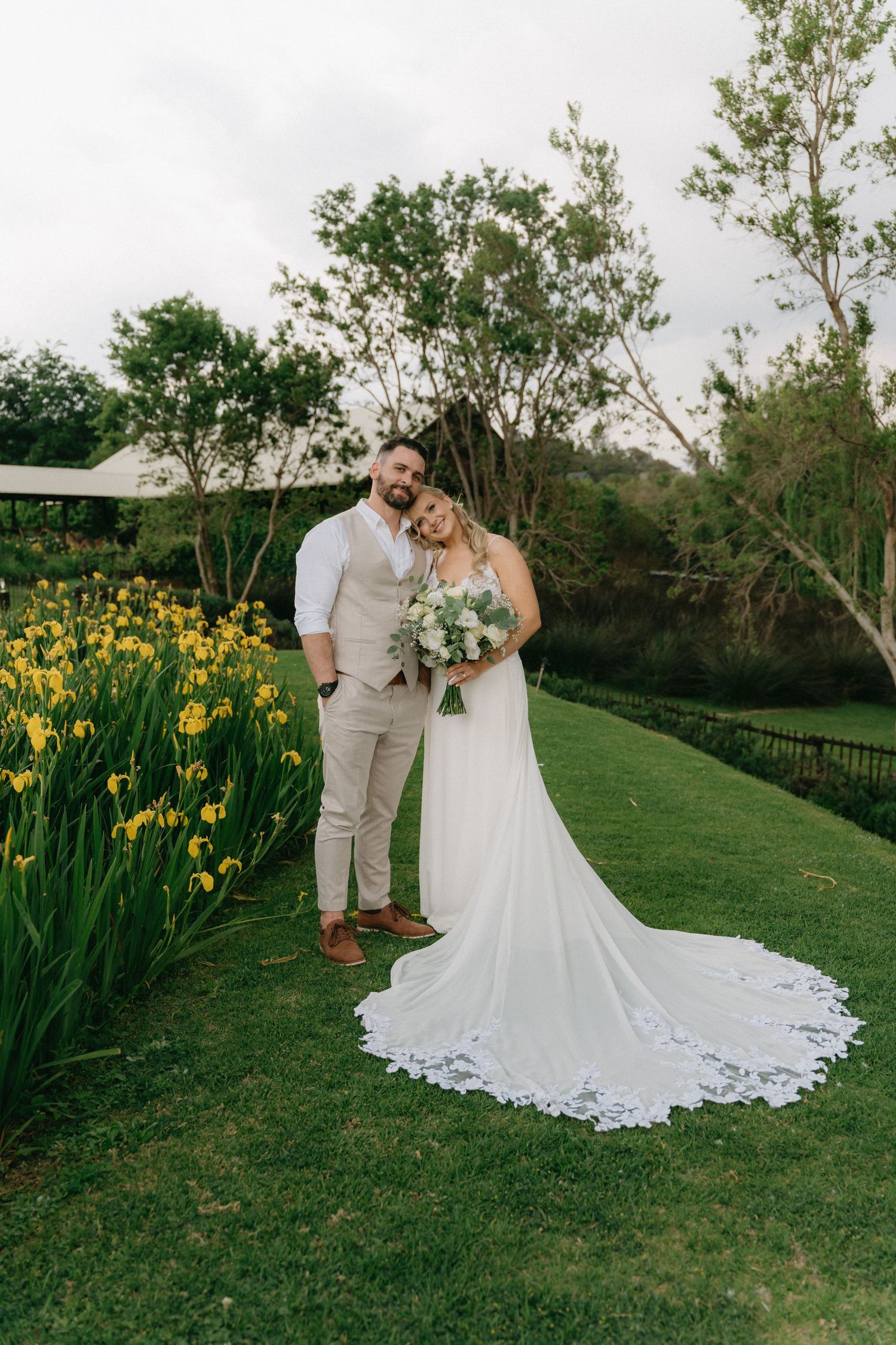 A bride in a lace wedding gown poses with her partner near blooming yellow daffodils.