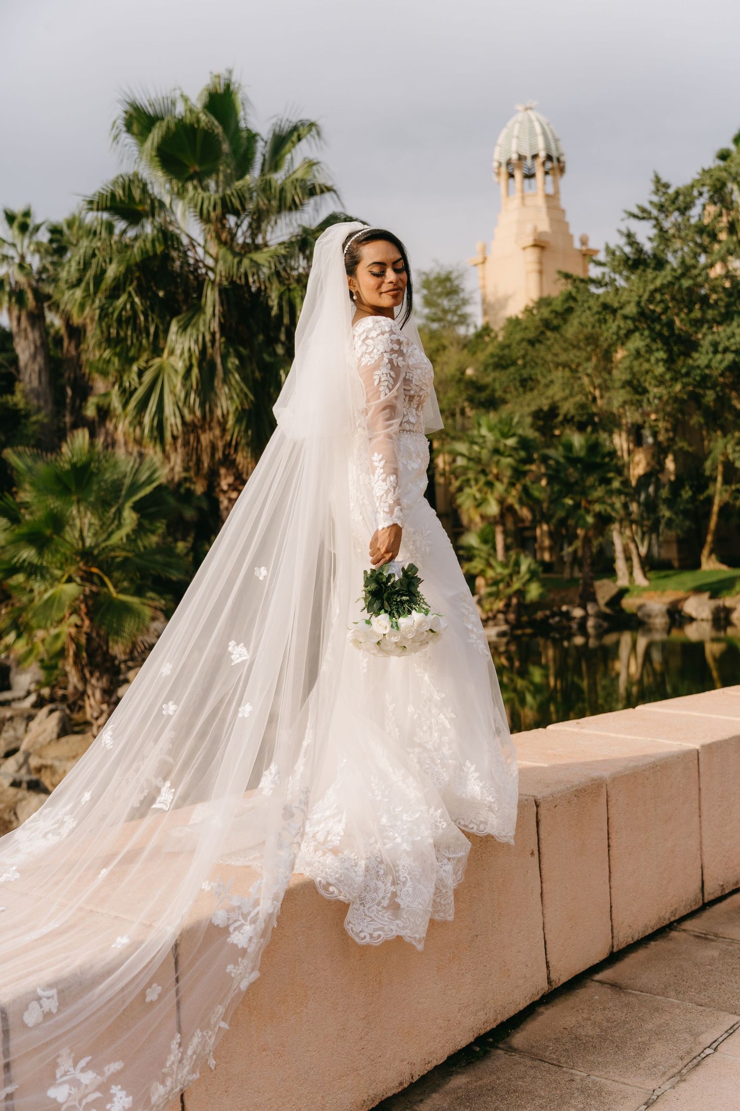 A flowing white lace wedding dress train cascades over beige stone rails at a palm-lined venue.