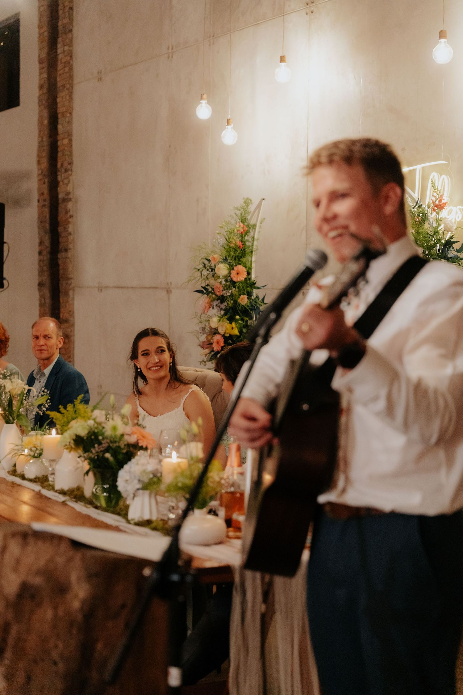 Wedding guests share laughter and joy during reception speeches in an elegant venue with hanging lights and floral decor.