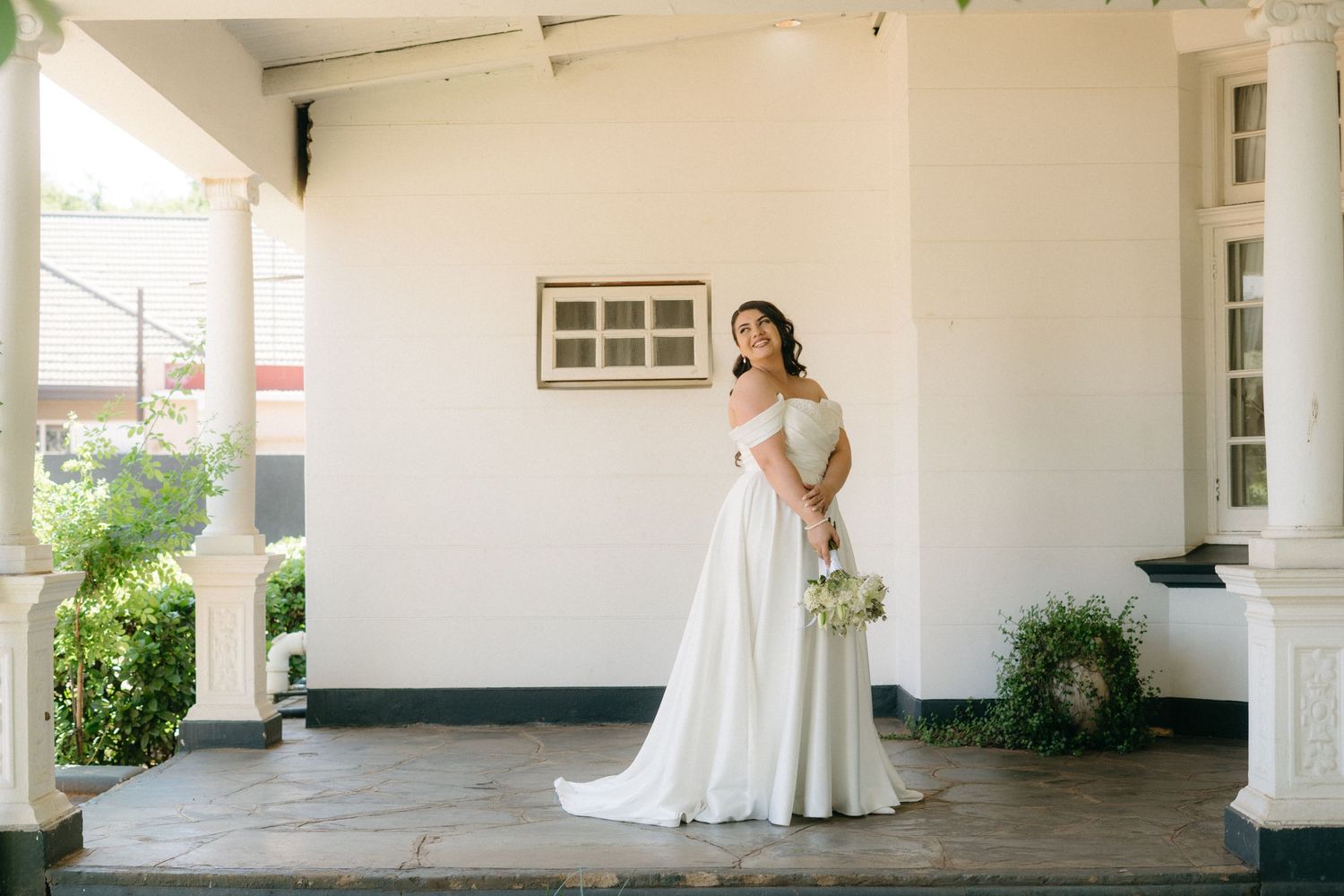 Elegant figure in a flowing white wedding gown stands gracefully on a bright white porch with classic columns and greenery.
