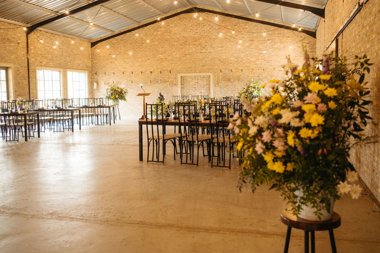 Rustic indoor wedding venue with exposed brick walls, string lights and yellow floral arrangements.