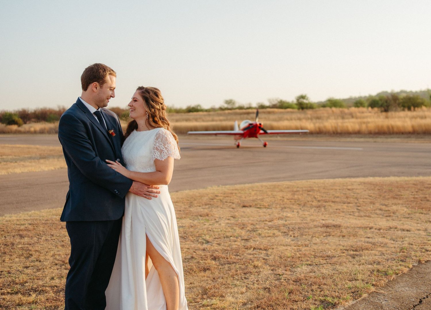 Newlyweds embrace on an airfield at sunset with a small red airplane in the background.