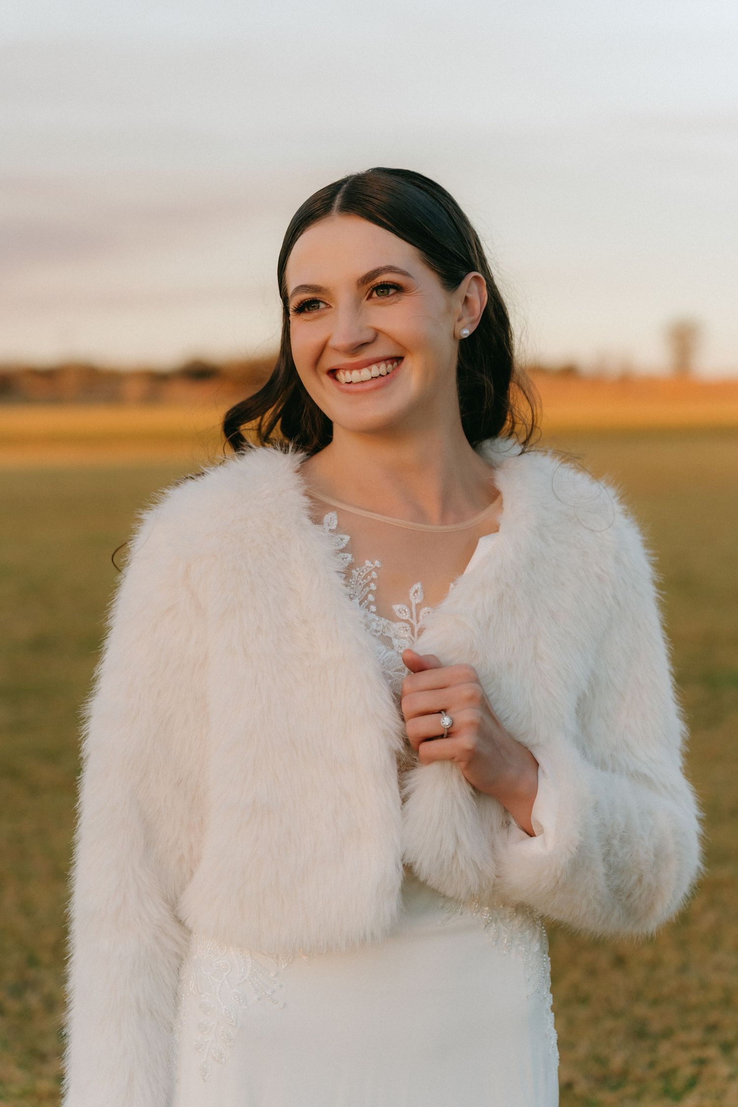 A bride wearing a white fur coat poses in a golden field during a winter sunset.